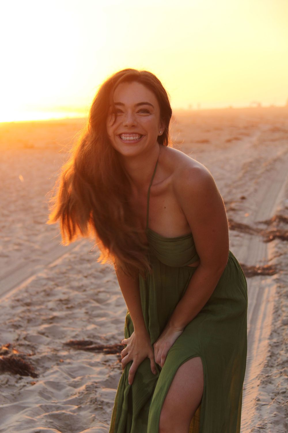 Woman in green dress laughs joyfully on beach at golden sunset.