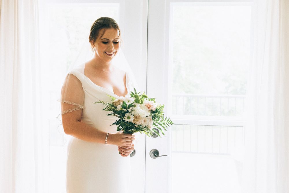 A bride in a classic white wedding dress holds a delicate bouquet of white and blush flowers by a bright window.