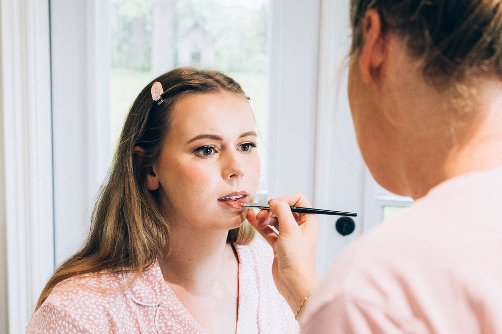 Makeup artist applies lipstick on a client wearing a pink robe in natural window lighting.