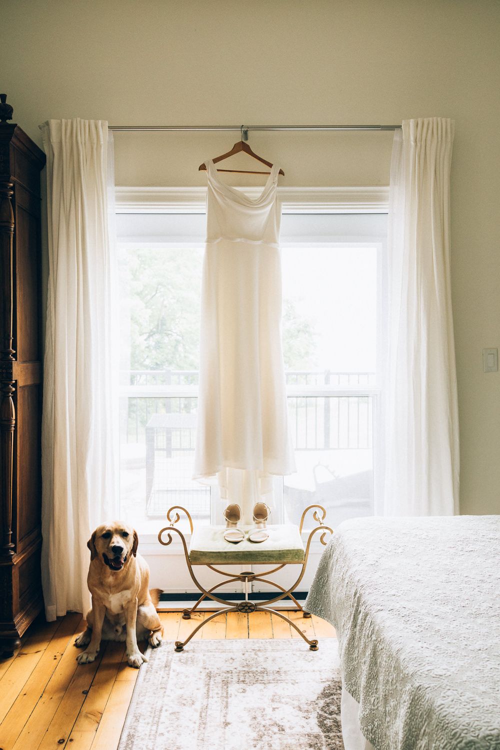 White wedding dress hanging in window with cream curtains and golden shoes, while dog sits nearby.