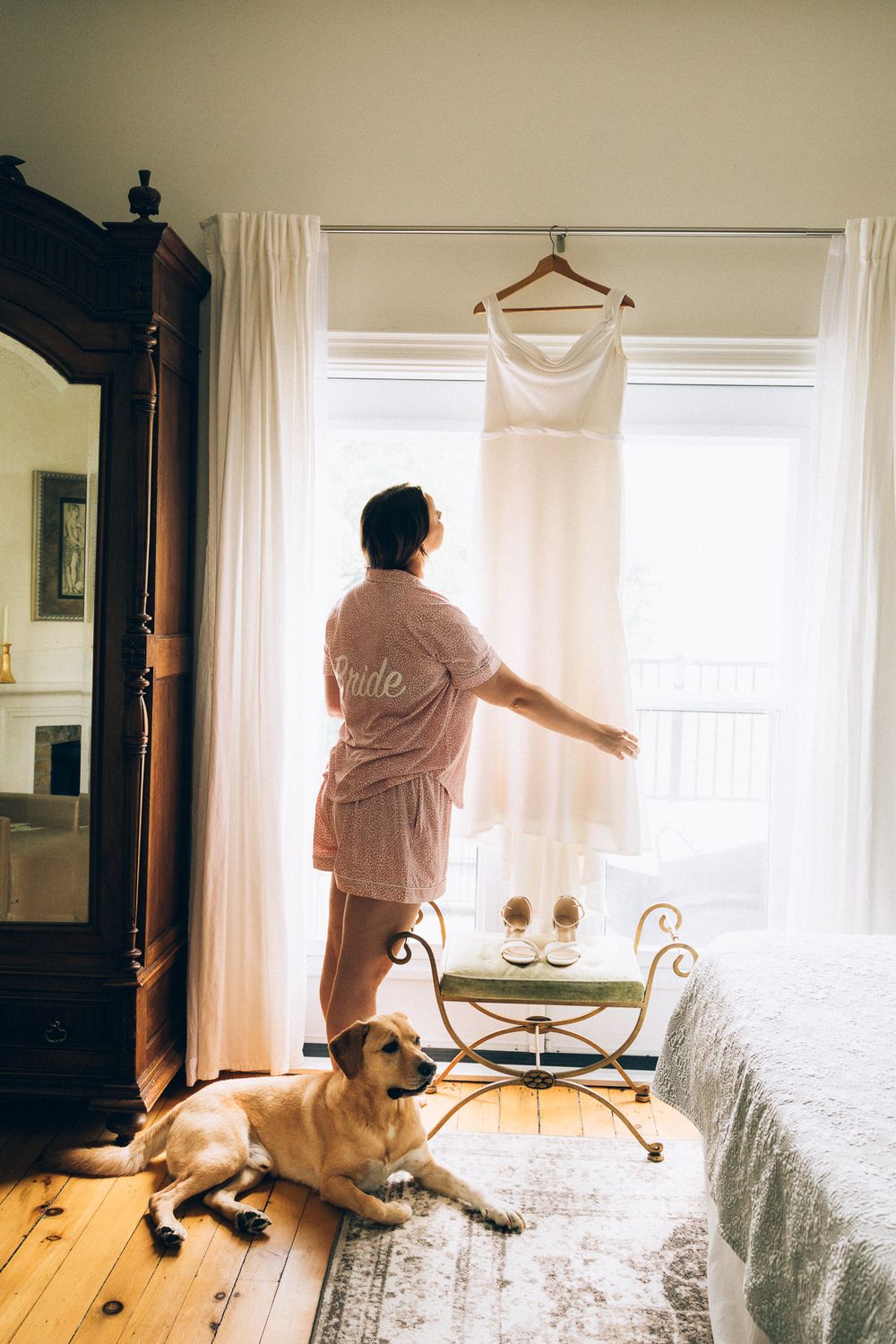 Person in pink robe reaches for wedding dress while dog rests on wooden floor by window.