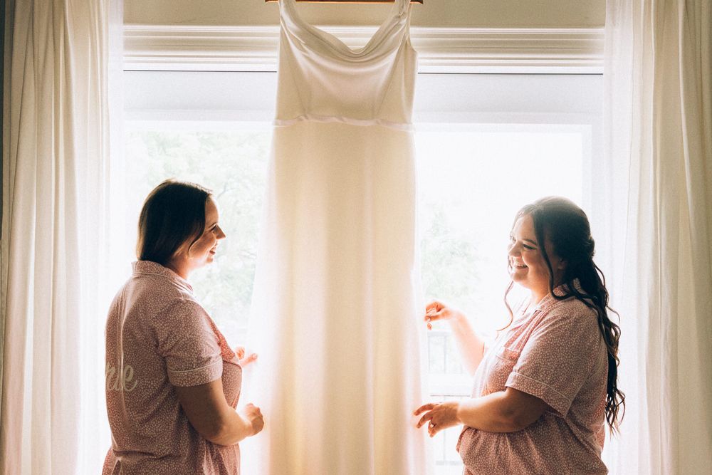Two people in robes discuss wedding dress hanging in bright window.