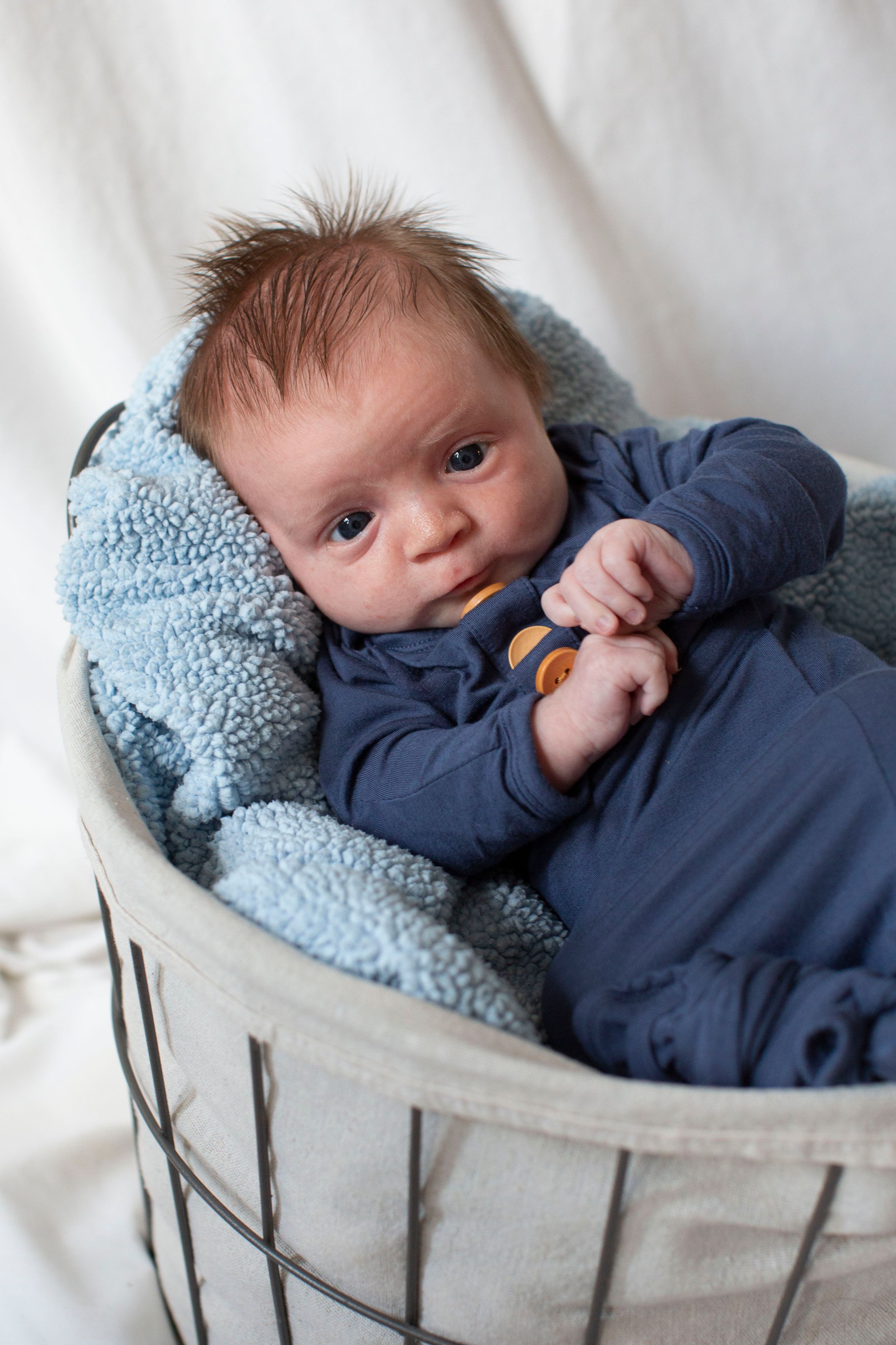 Newborn baby in blue onesie resting in a wire basket lined with soft blue blanket.