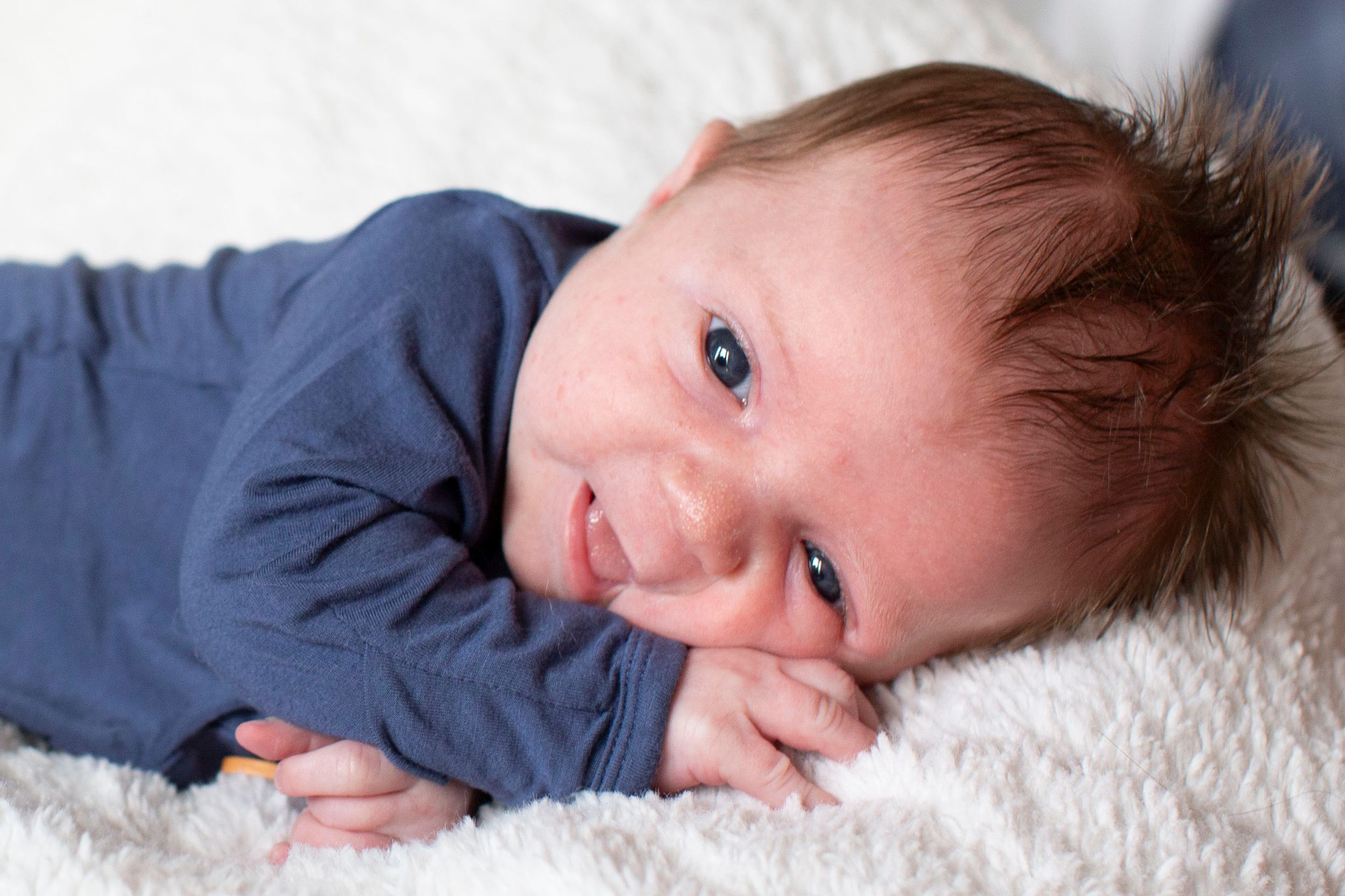 Adorable newborn baby smiling while lying on a fluffy white blanket in blue outfit.