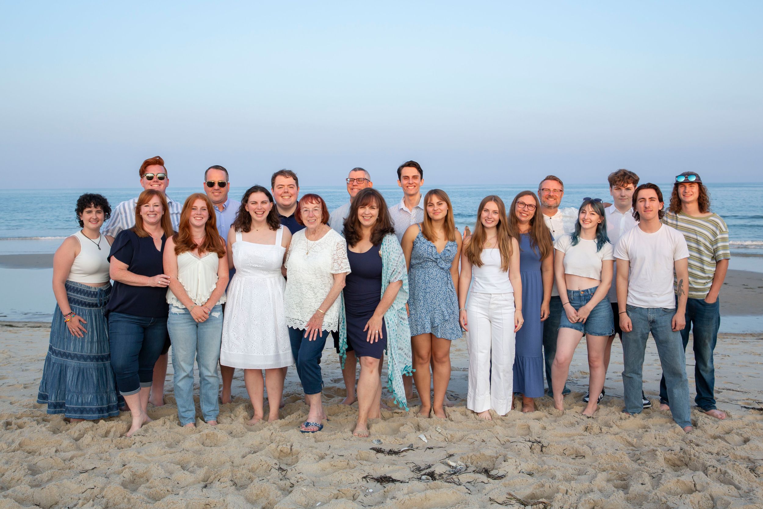 Large family group portrait on a sandy beach at dusk, casually dressed in coordinating blue and white.