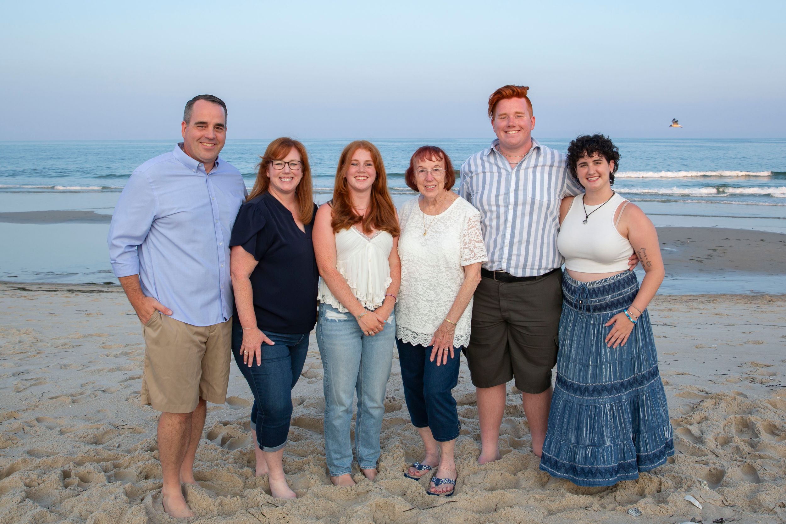 Family of six smiling together on a beach at sunset in coordinating casual outfits.