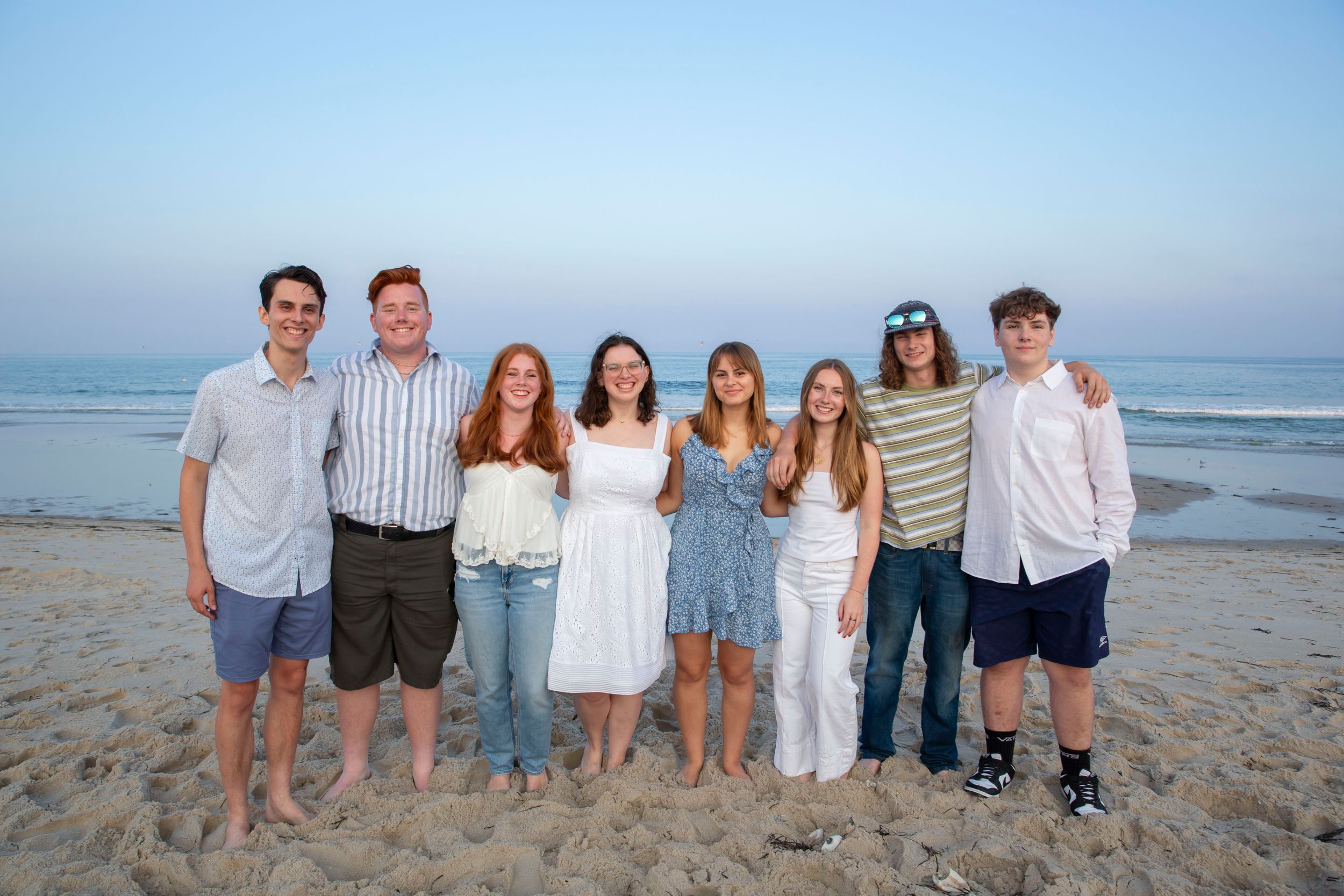 Group of eight young adults posing together on a beach at dusk in coordinating outfits.