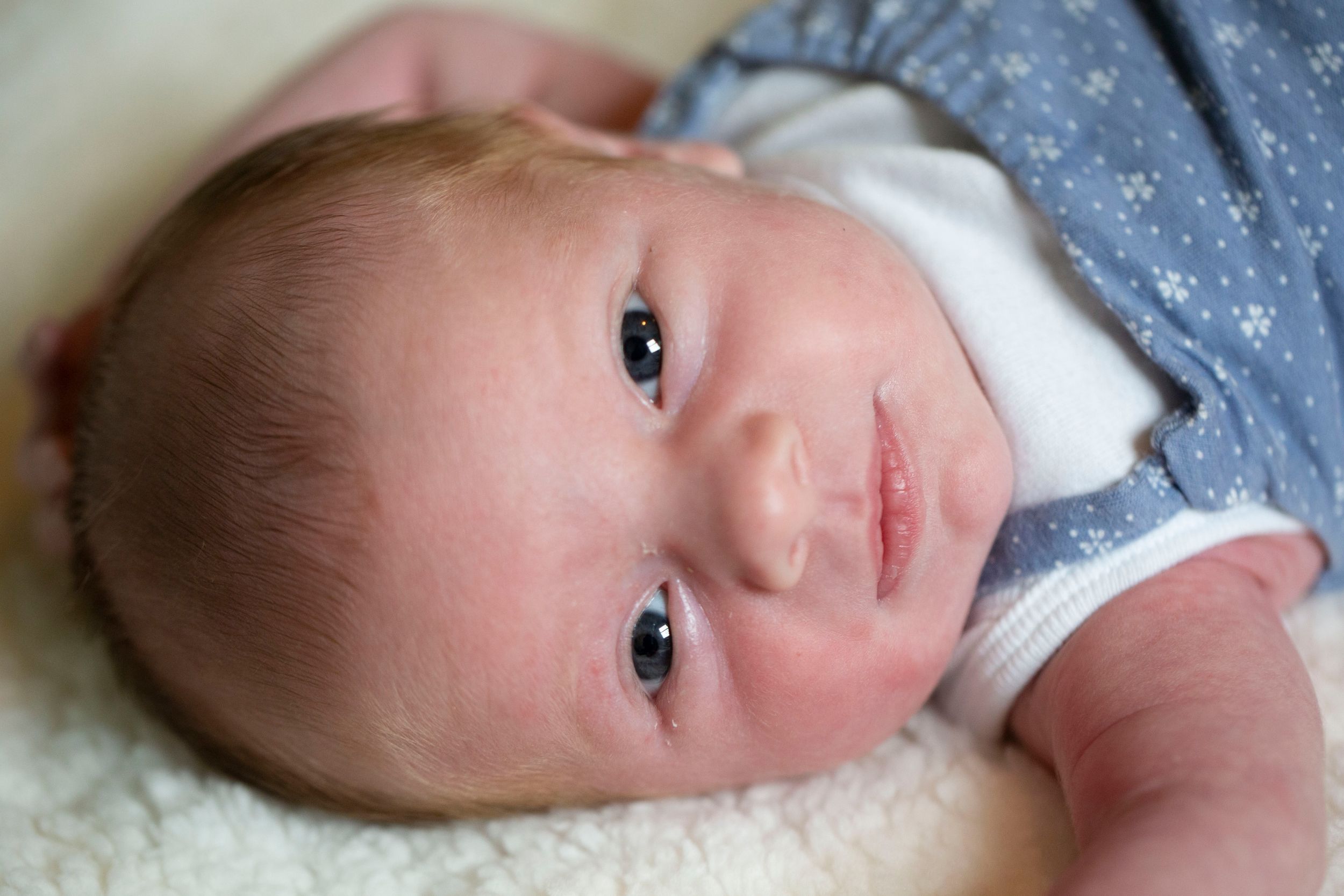 Close-up portrait of a newborn baby in a blue outfit lying on a soft white blanket.