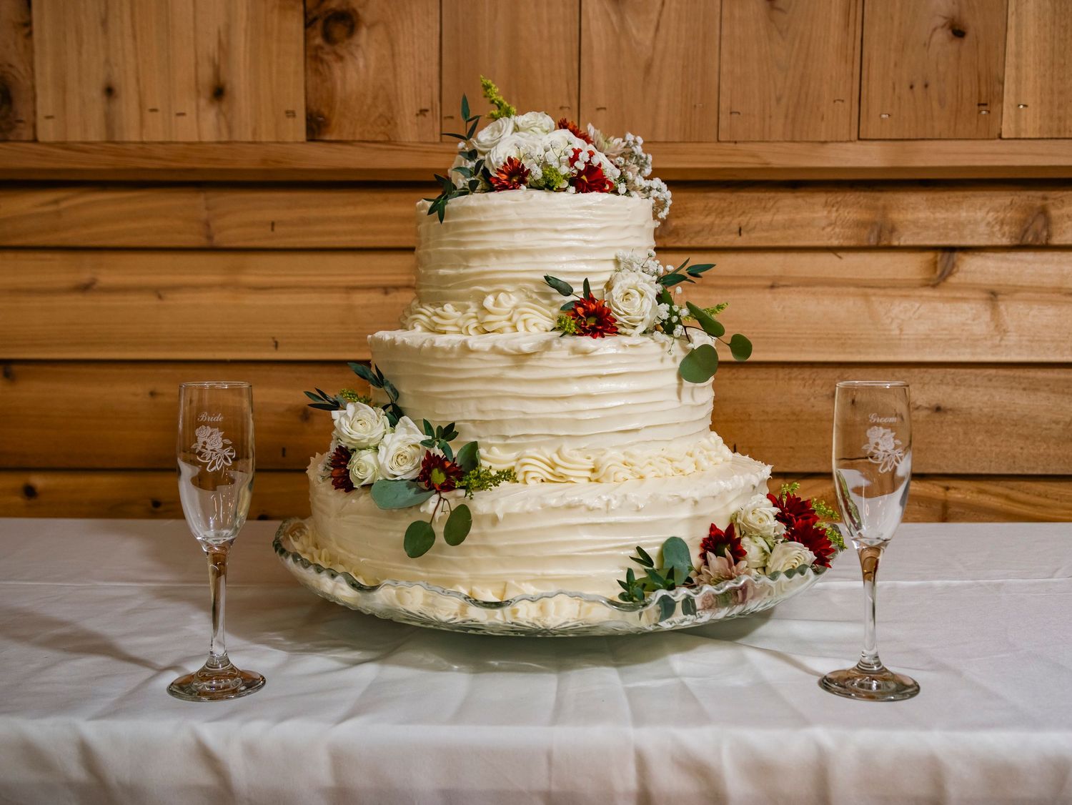 Three-tiered white wedding cake decorated with red flowers and greenery on white tablecloth.
