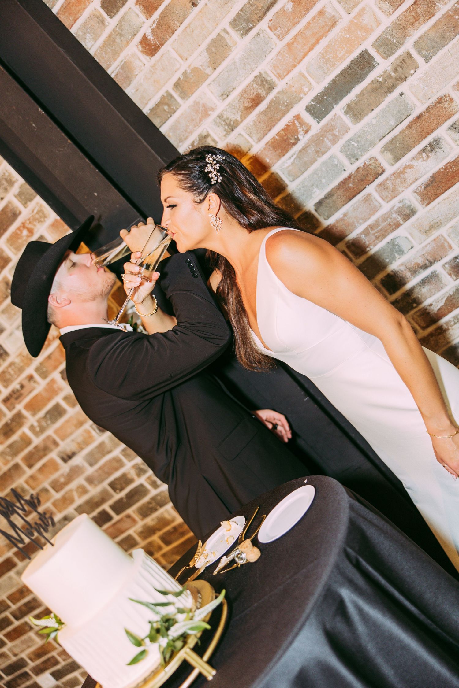 Wedding couple sharing playful cake-cutting moment against brick wall backdrop.
