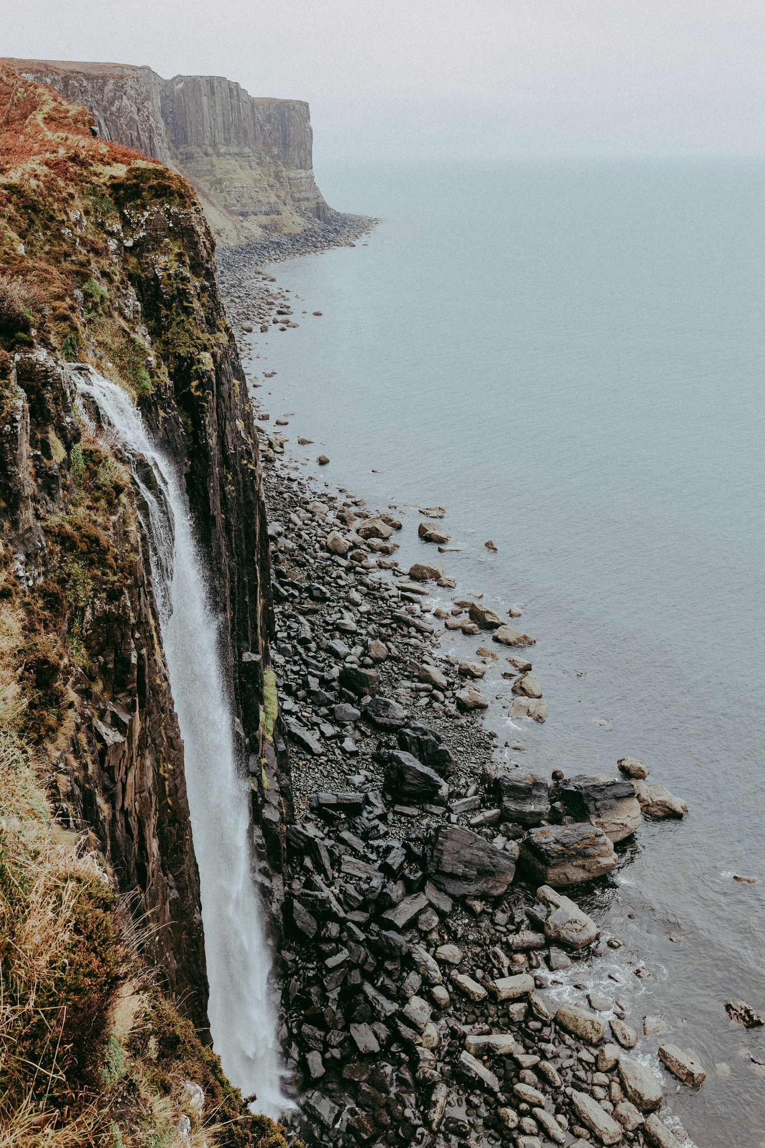 A dramatic waterfall cascades down rugged coastal cliffs onto a rocky shoreline with calm grey sea in the background. Popp Photography