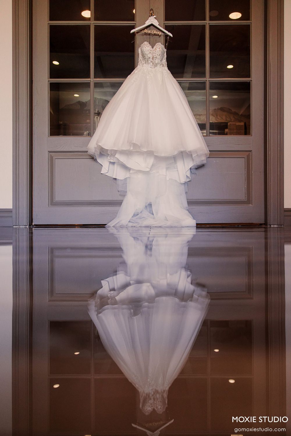 Elegant white wedding dress displayed in front of dark reflective surface with dramatic lighting and mirrored reflection.
