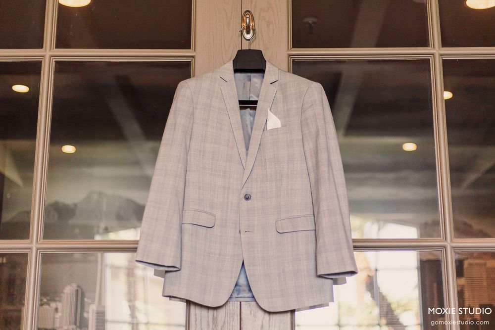 Light gray and white suits displayed on mannequins in a retail store window with pendant lighting above.