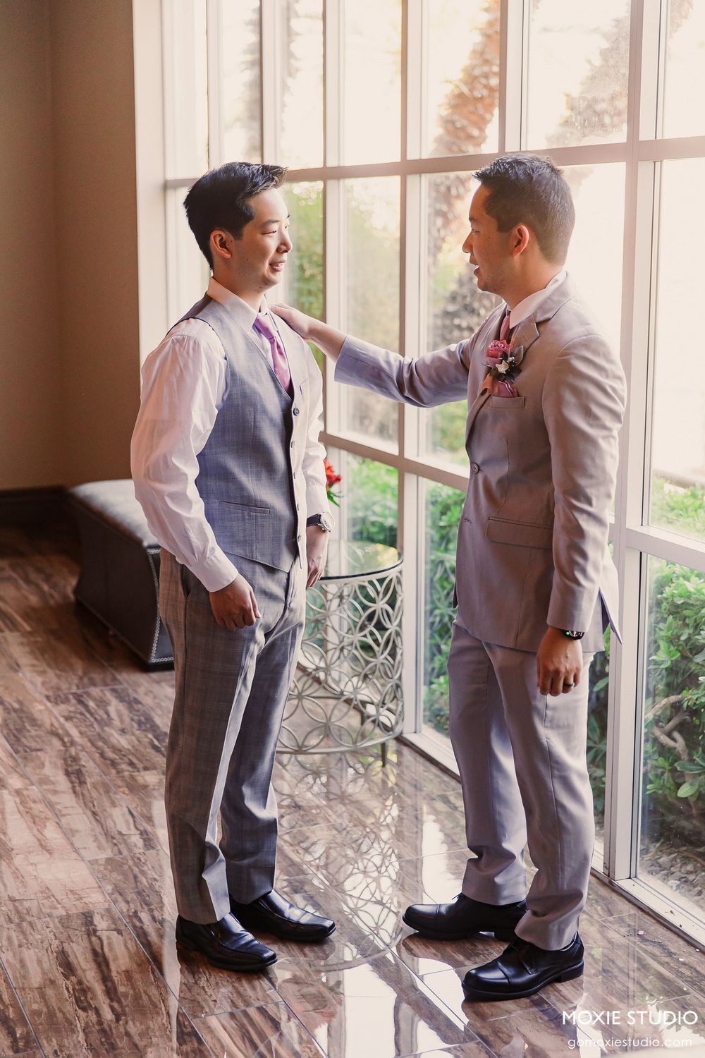 Two young boys in gray suits stand together near a bright window in a formal setting.