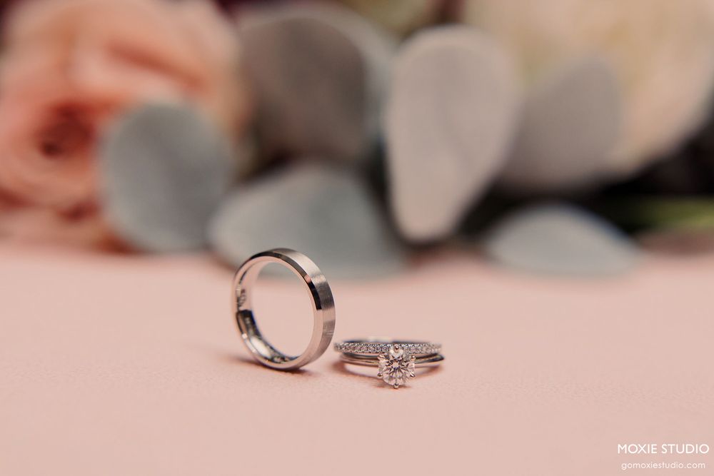 Wedding rings displayed on a soft pink surface with blurred eucalyptus leaves in the background.