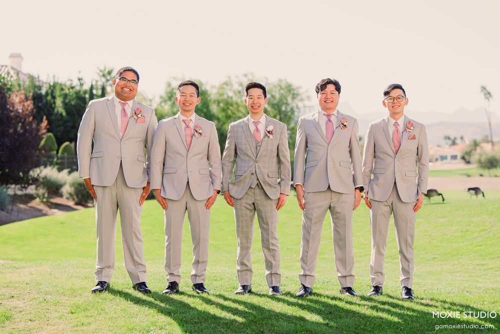 Groomsmen in light gray suits with pink ties stand in a row on a grassy lawn at an outdoor wedding venue.
