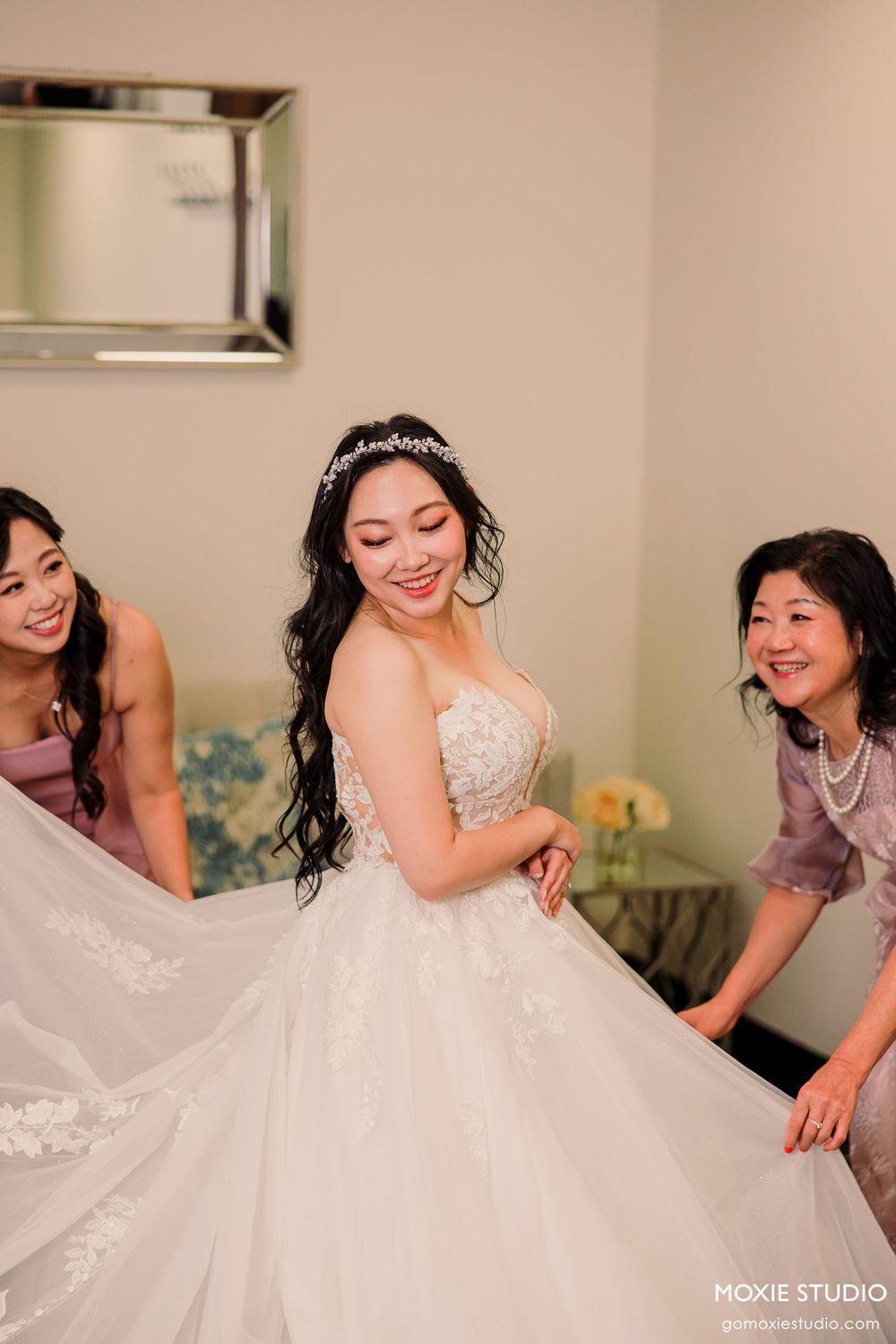 Bridesmaids in pink dresses assist with fluffing and arranging a bride's voluminous white wedding gown.