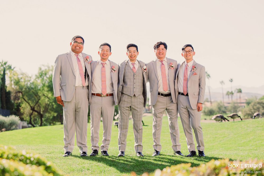 Wedding party members in coordinated gray suits pose together on a sunny outdoor lawn.