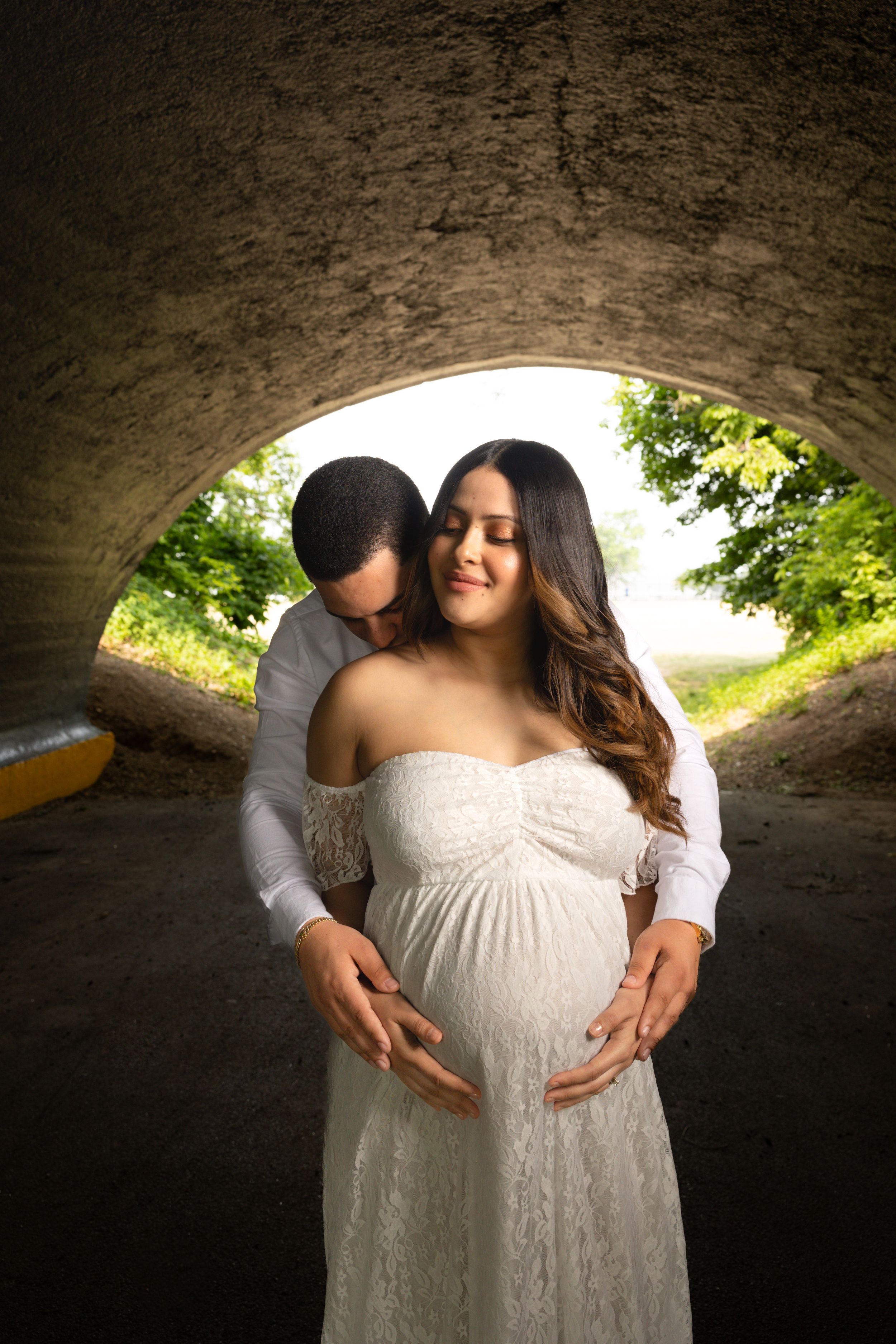 Intimate maternity photo under stone arch tunnel with couple embracing in white lace dress and soft natural lighting.