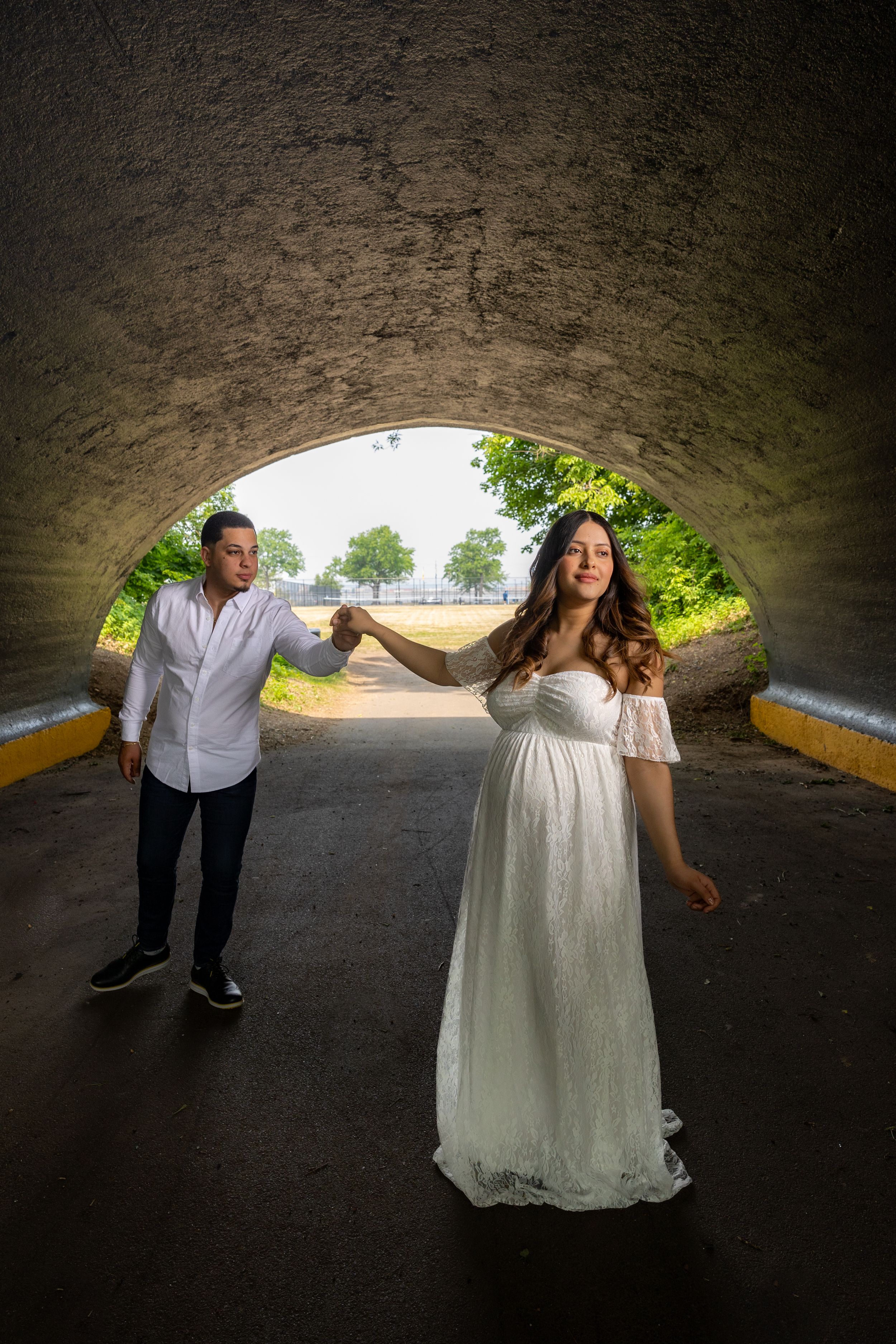 A couple in a romantic tunnel pose with the bride wearing a white off-shoulder dress under a stone archway posing for a maternity shoot .