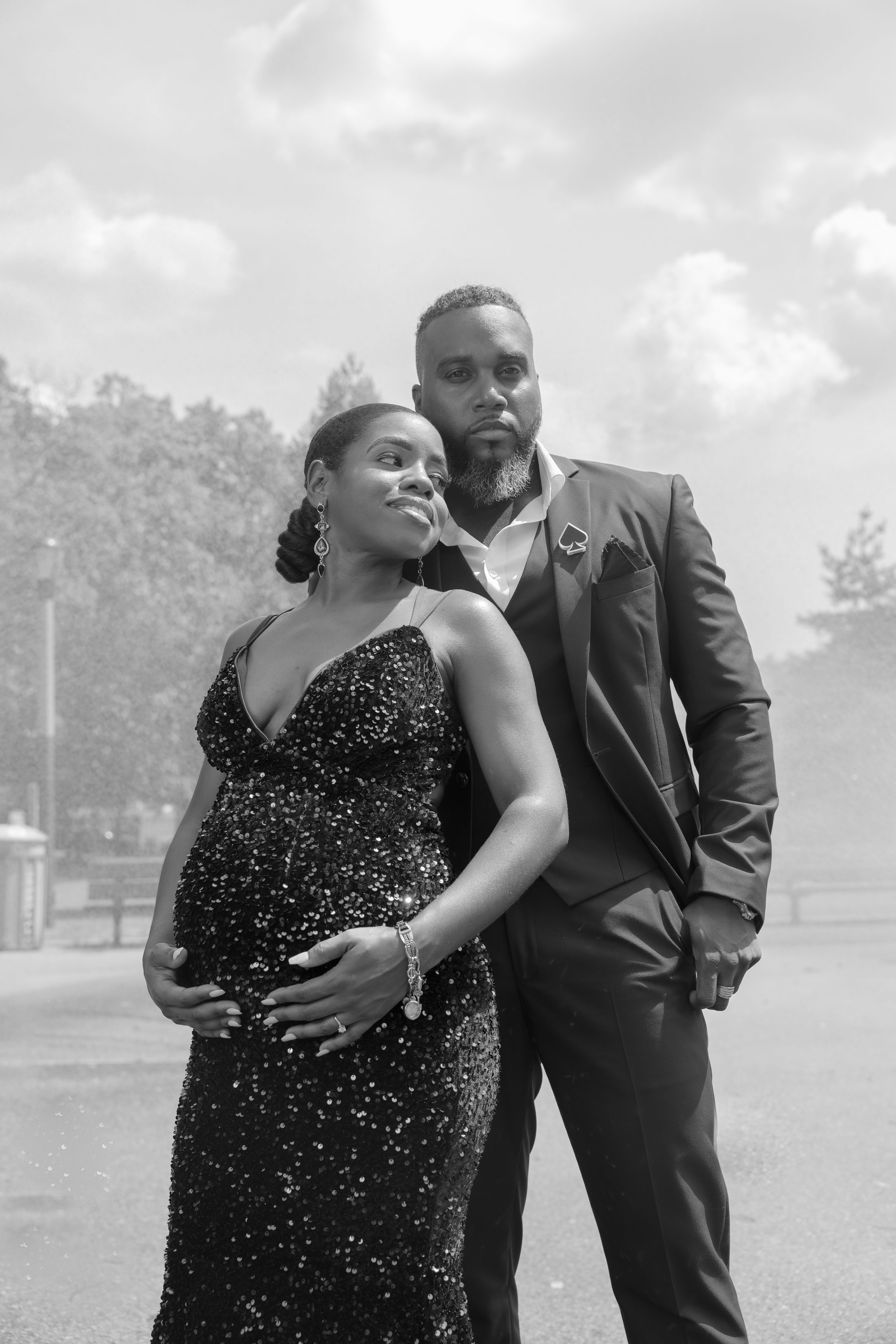 A couple poses outdoor for a maternity shoot, in formal attire, with the woman wearing a sparkly black gown and cradling her pregnant belly.