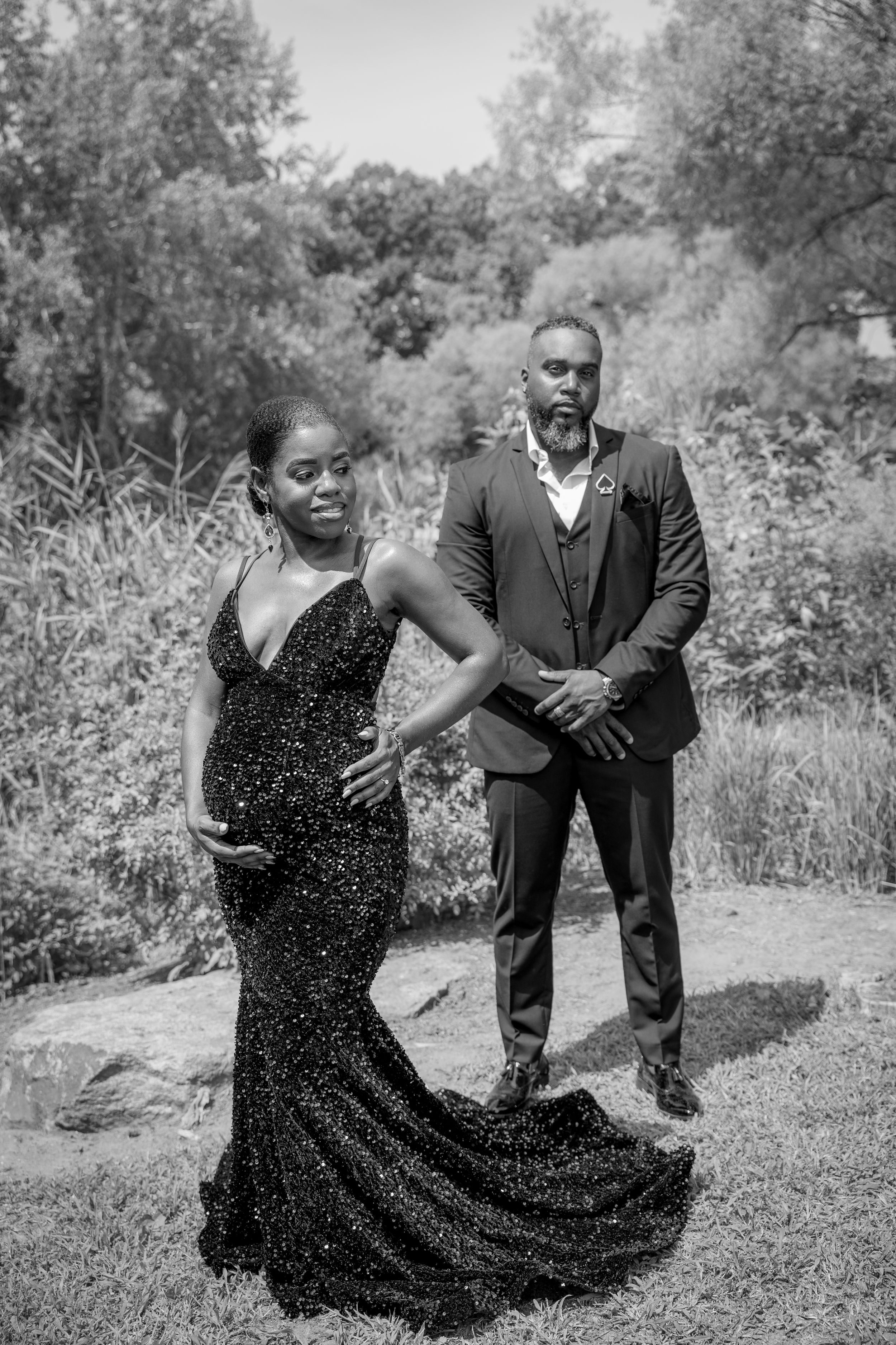 A romantic couple poses outdoors for a maternity shoot, with her in a sparkly black gown and him in a sharp suit surrounded by greenery