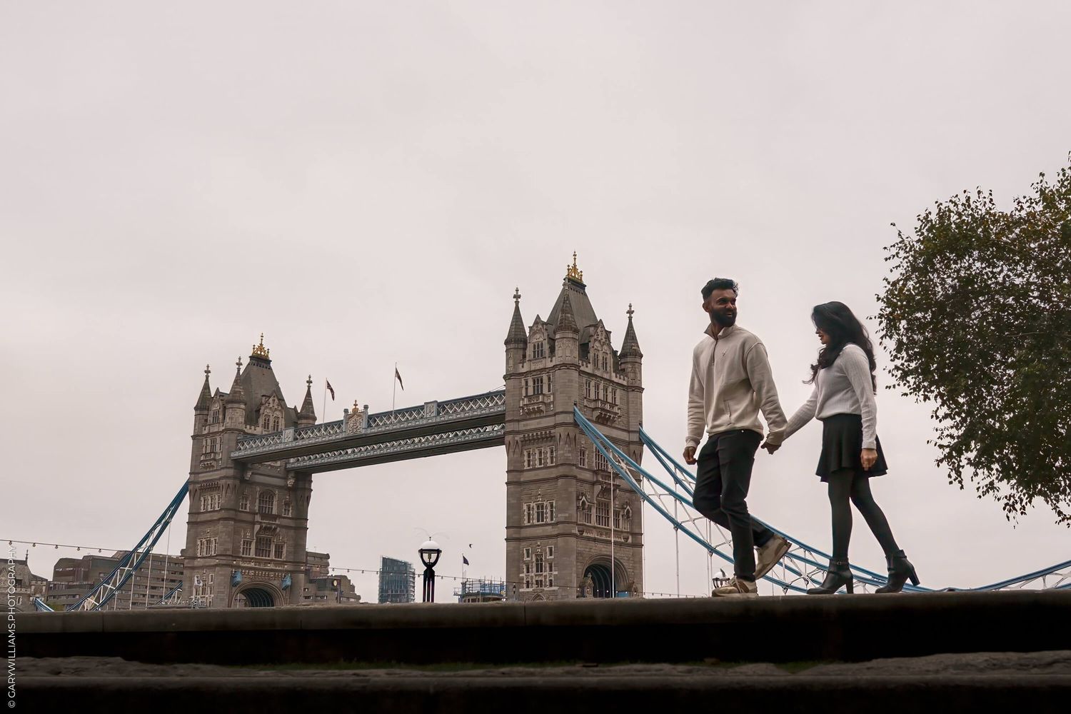 Couple walking along wall with Tower Bridge in background during overcast day in London.