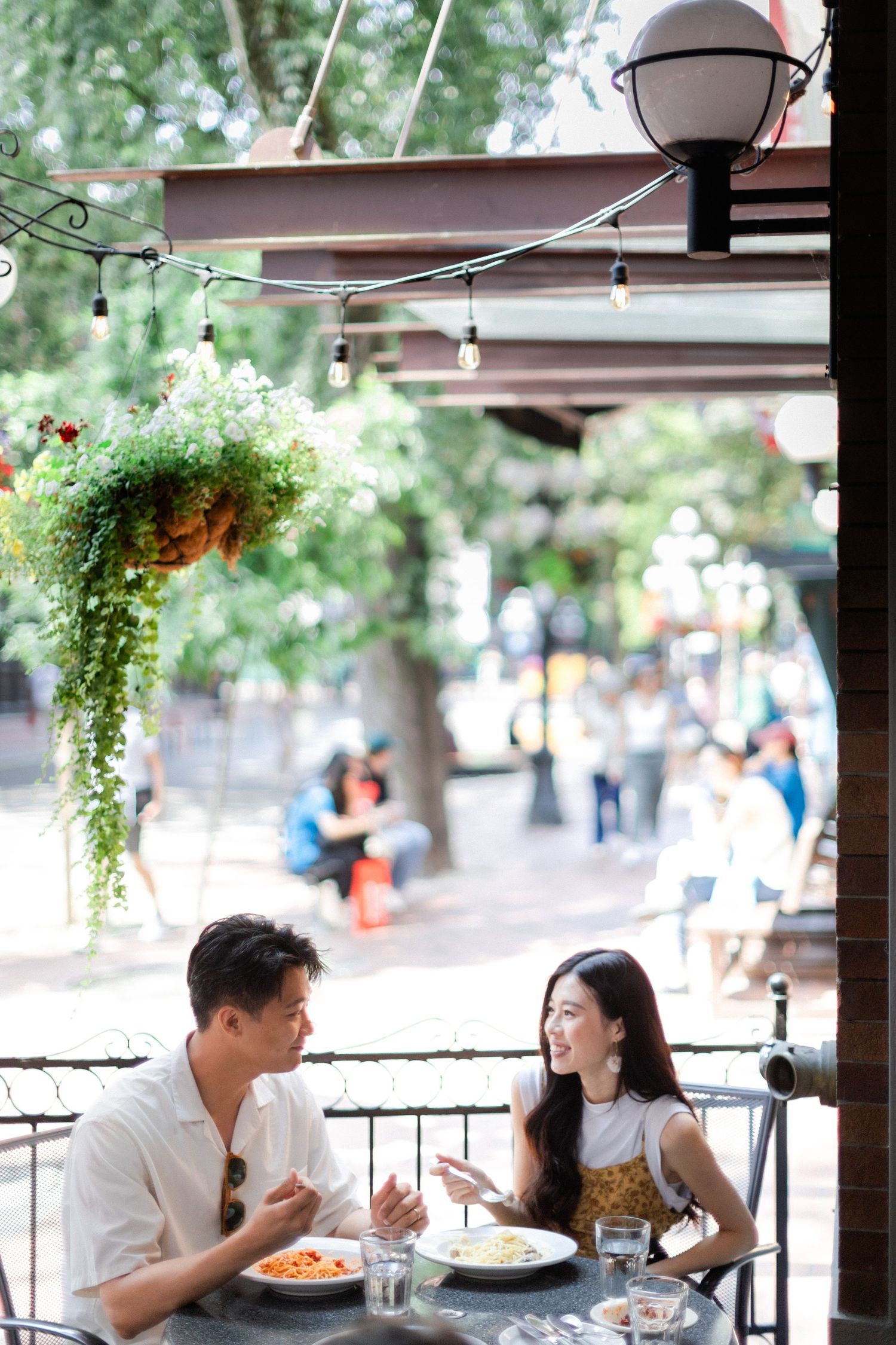 Couple enjoying a romantic outdoor lunch at a patio restaurant with hanging plants and string lights.