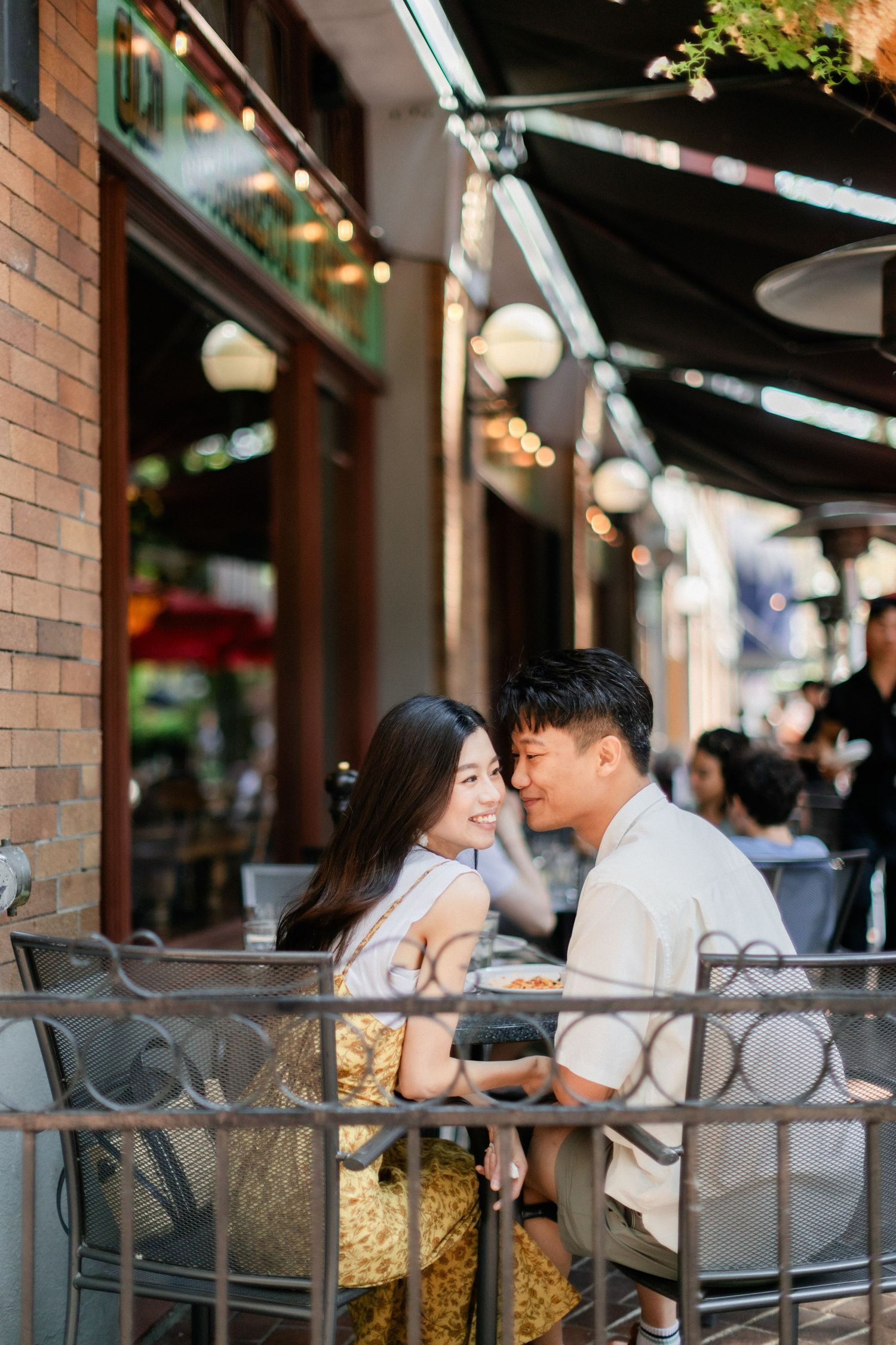 Young Asian couple sharing a romantic moment at an outdoor cafe patio with warm bokeh lights.