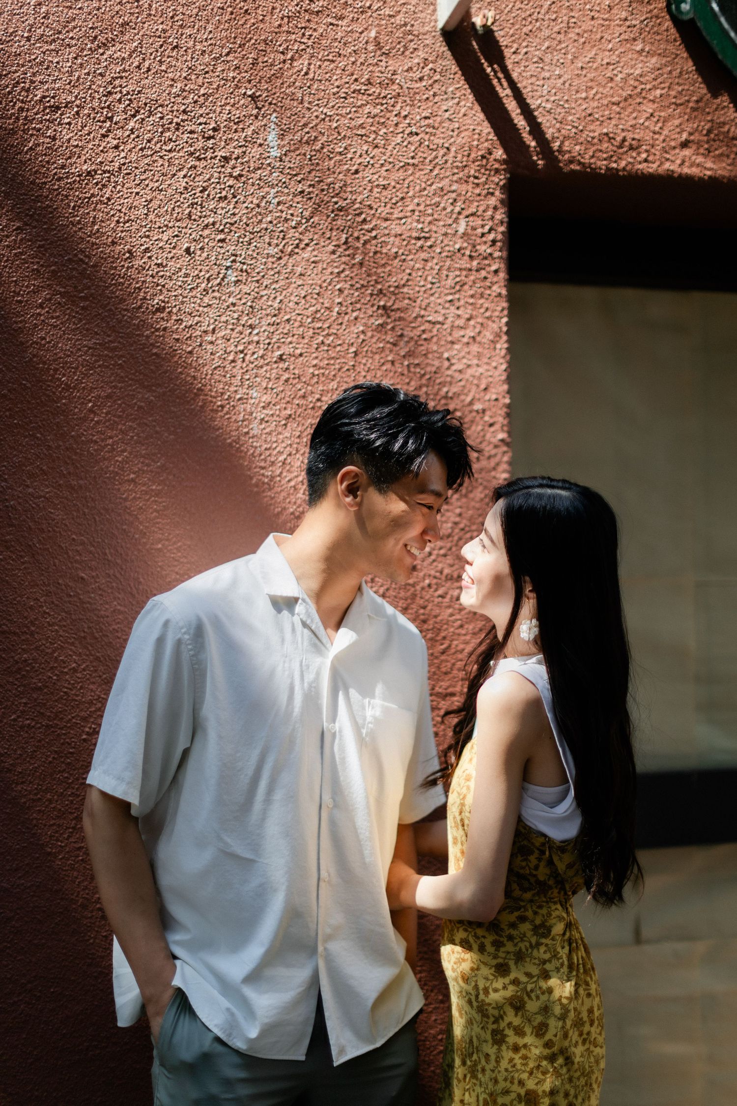 Young Asian couple sharing an intimate moment against a sunlit terracotta wall outdoors.