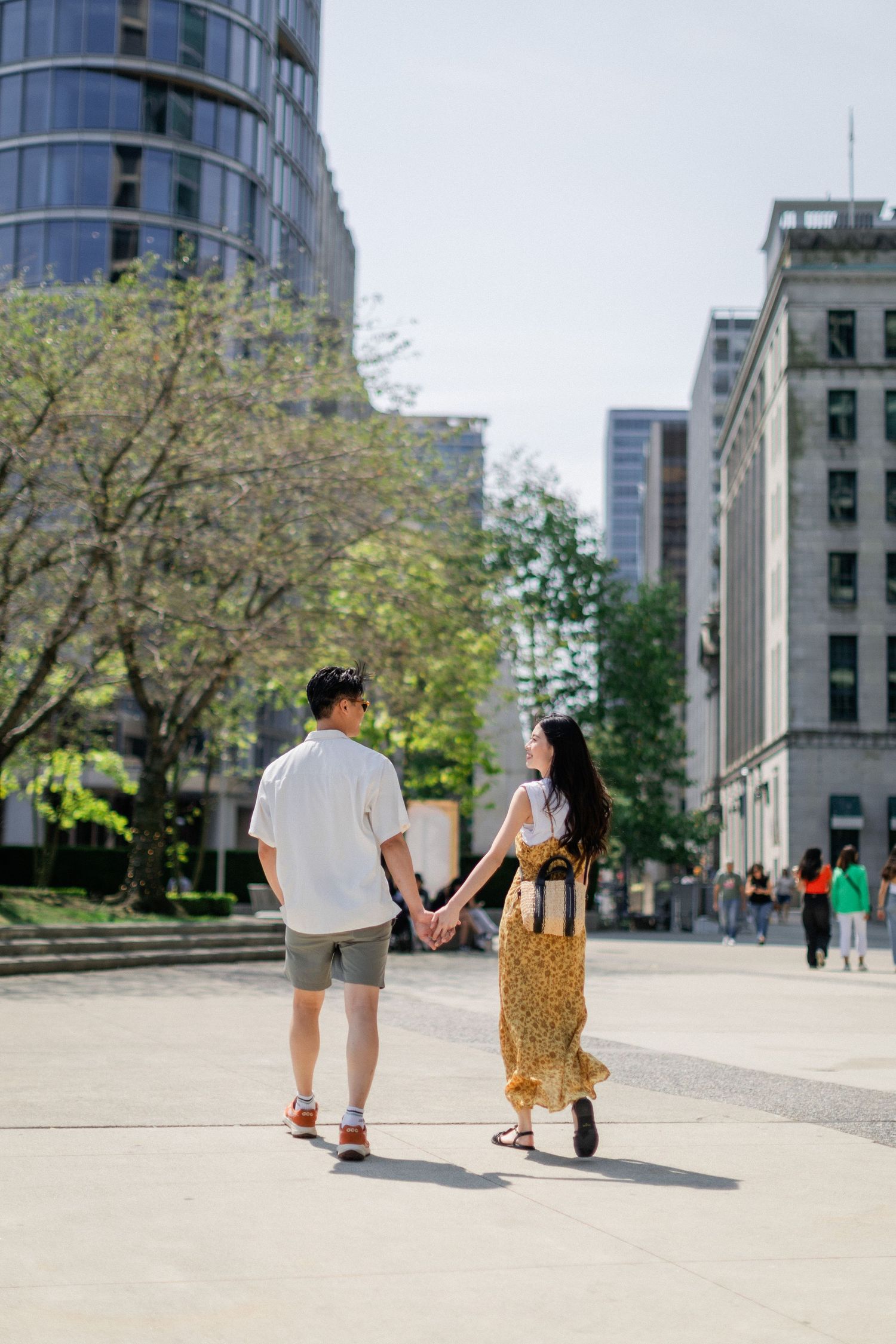Asian couple holding hands while walking through a sunny urban plaza surrounded by city buildings.