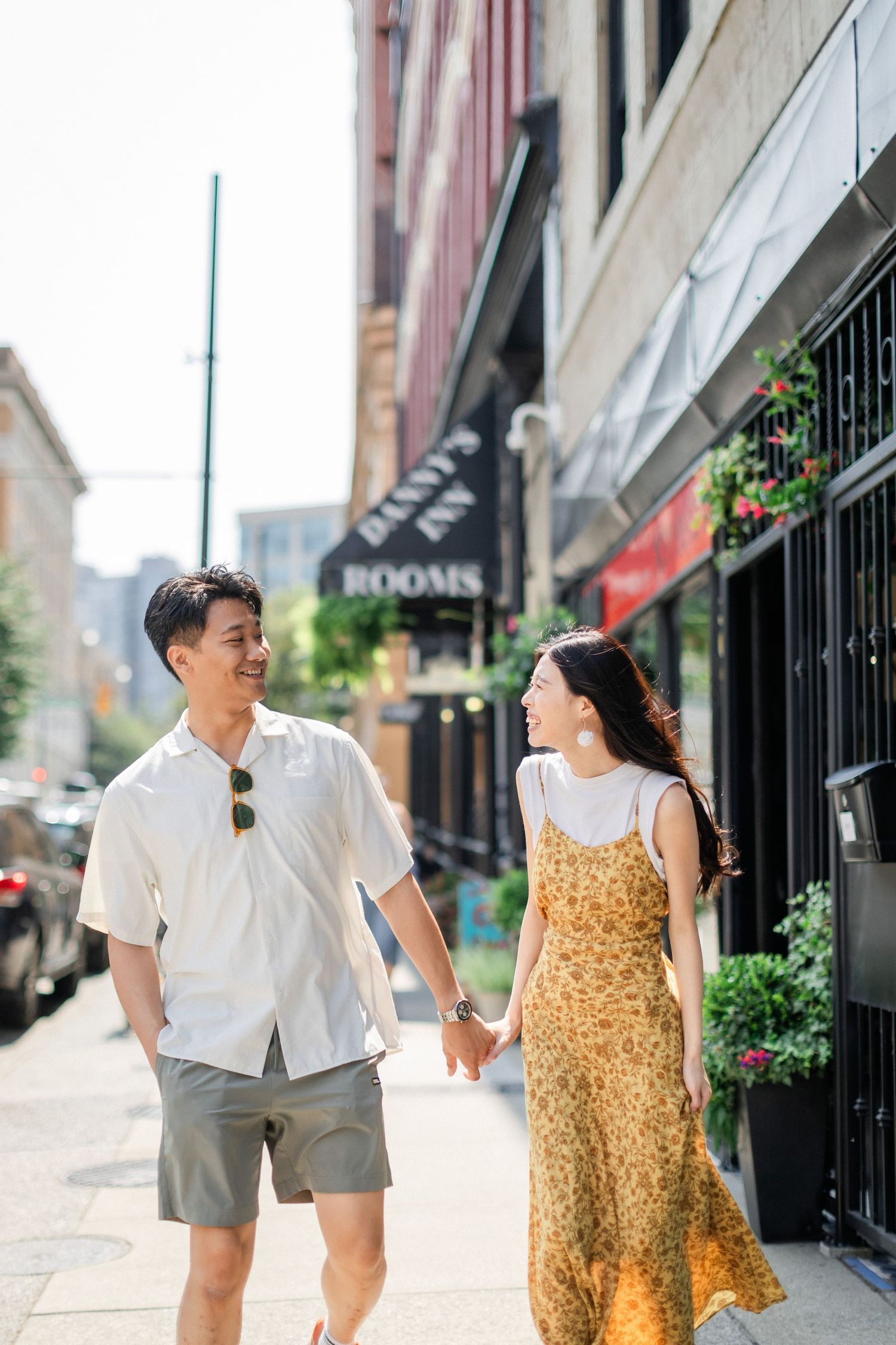 Smiling Asian couple holding hands while walking along a sunny city sidewalk past storefronts.