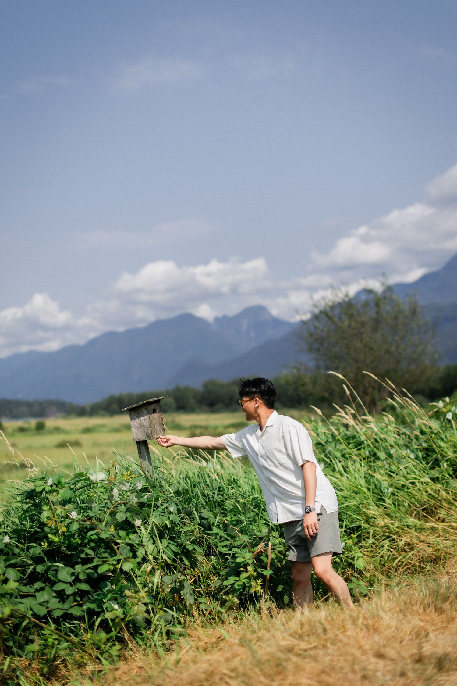 Young Asian man exploring a lush green meadow with mountains and blue sky in the background.