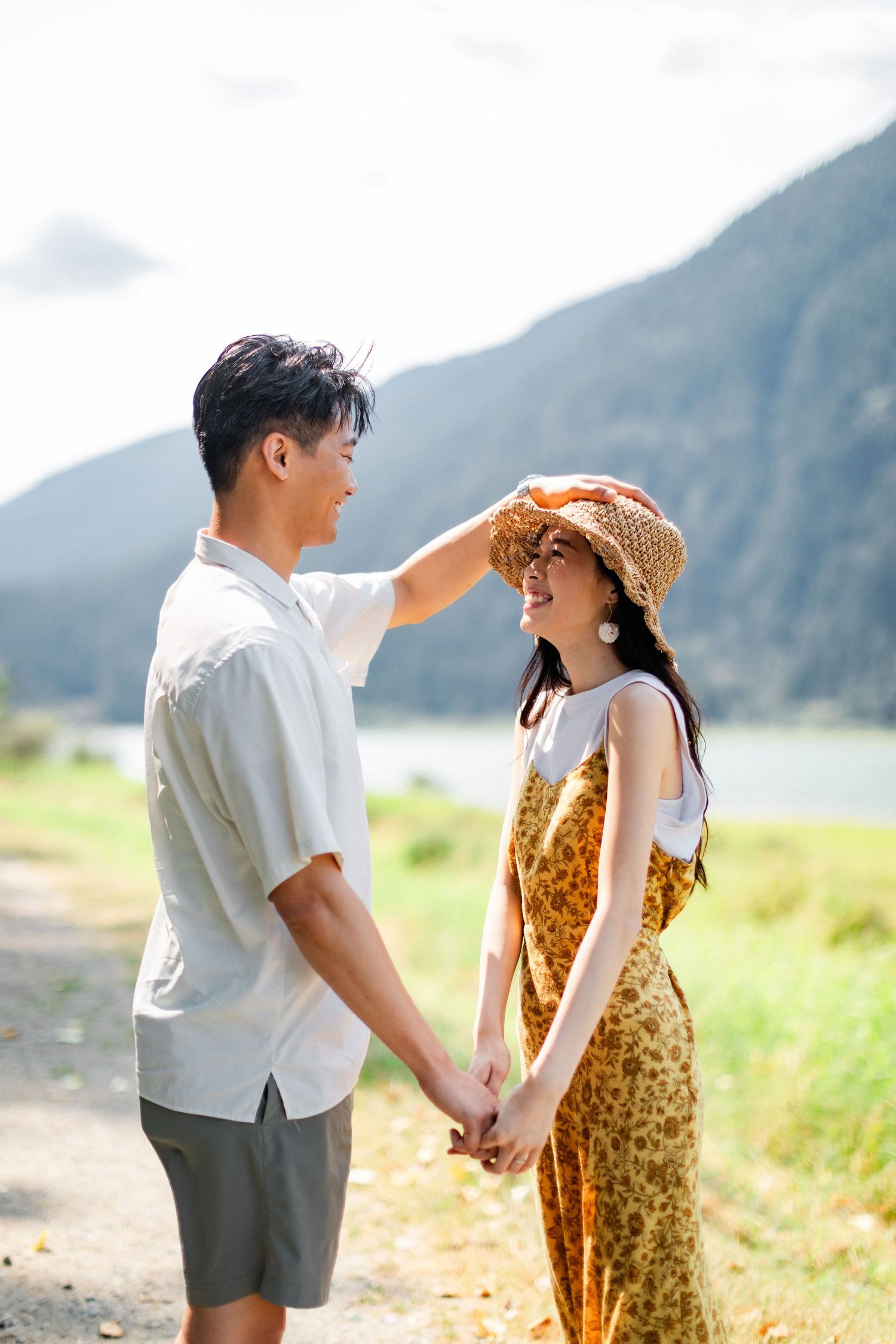 Asian couple holding hands outdoors with man placing a straw hat on womans head near mountains.