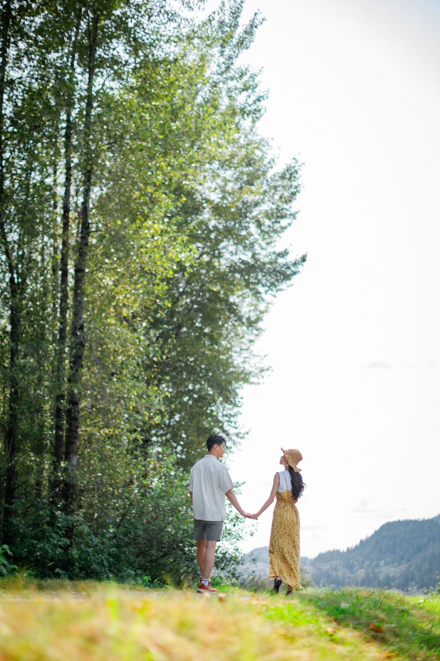 A couple holds hands while walking along a tree-lined path with mountains in the background.