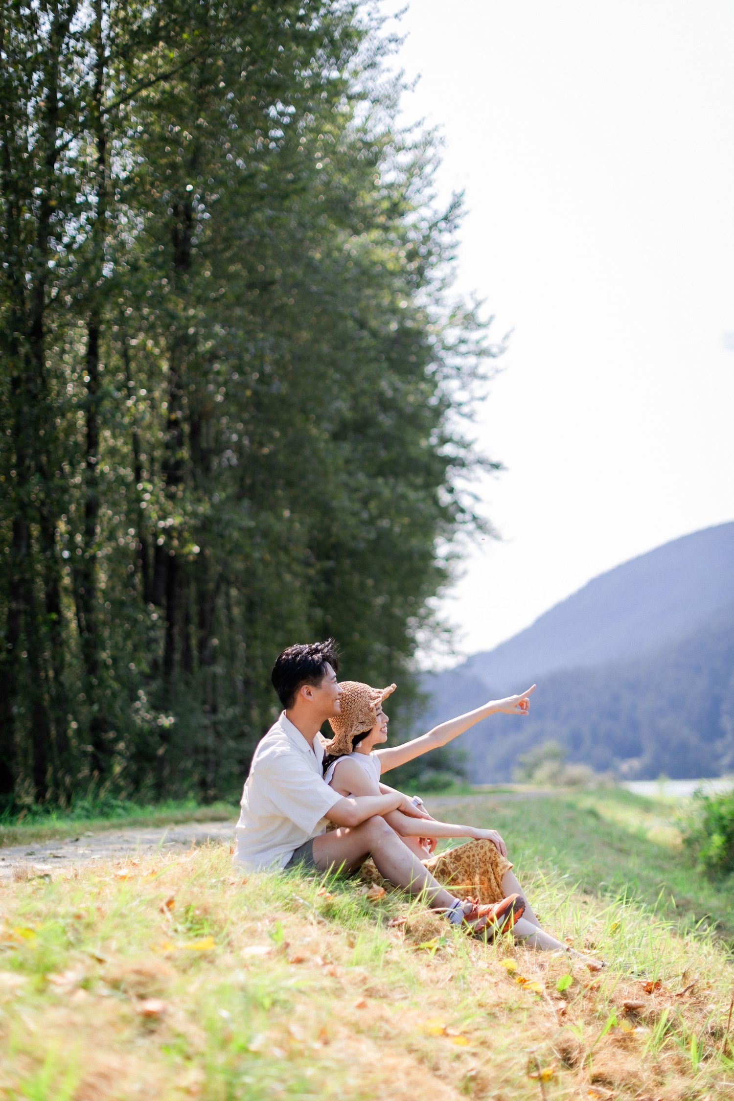 A couple sits on a grassy hillside, with the woman pointing toward distant mountains.