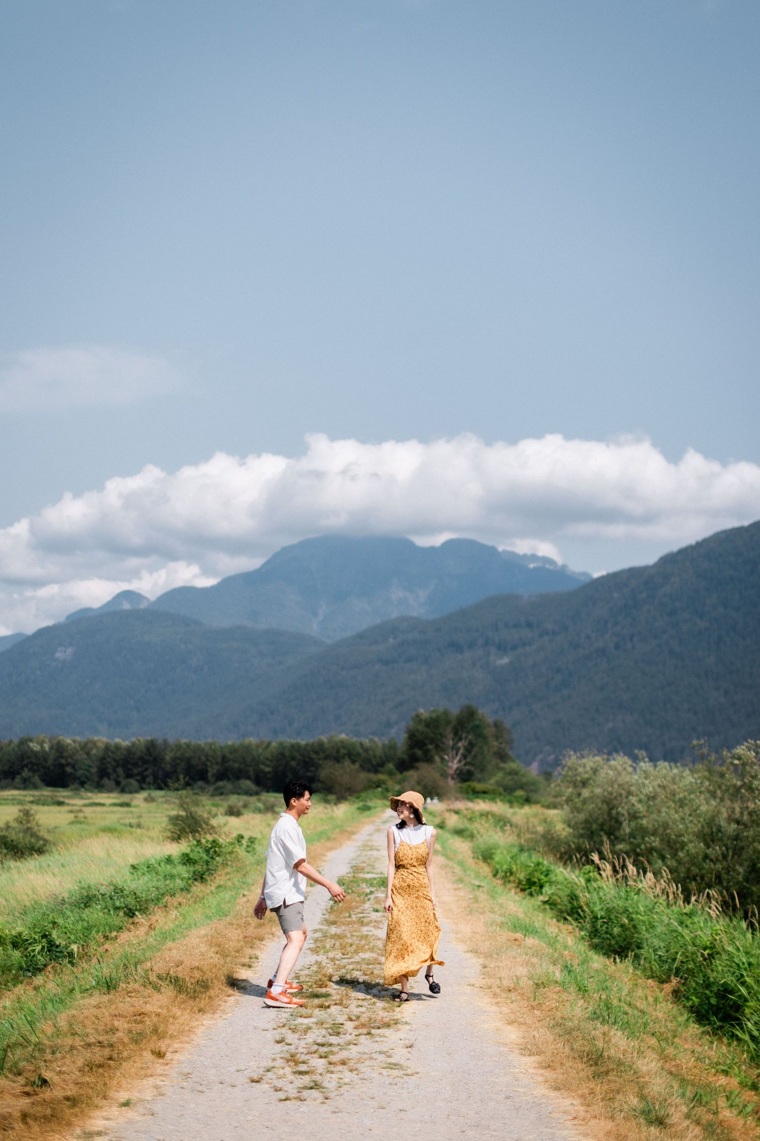A couple holds hands while walking down a rural path surrounded by green fields and mountains.