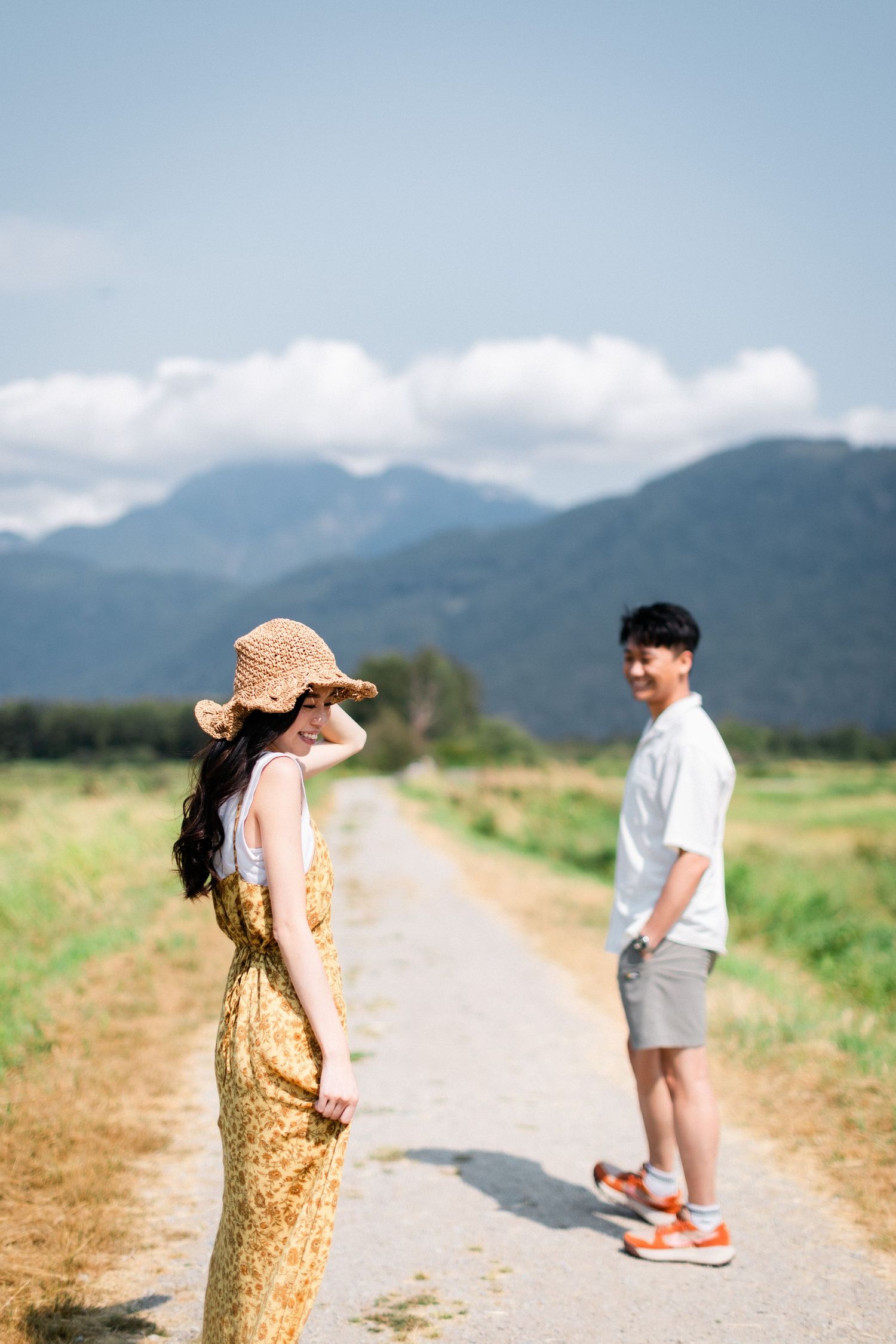 A woman in a yellow floral dress and straw hat smiles on a path as a man stands behind her.