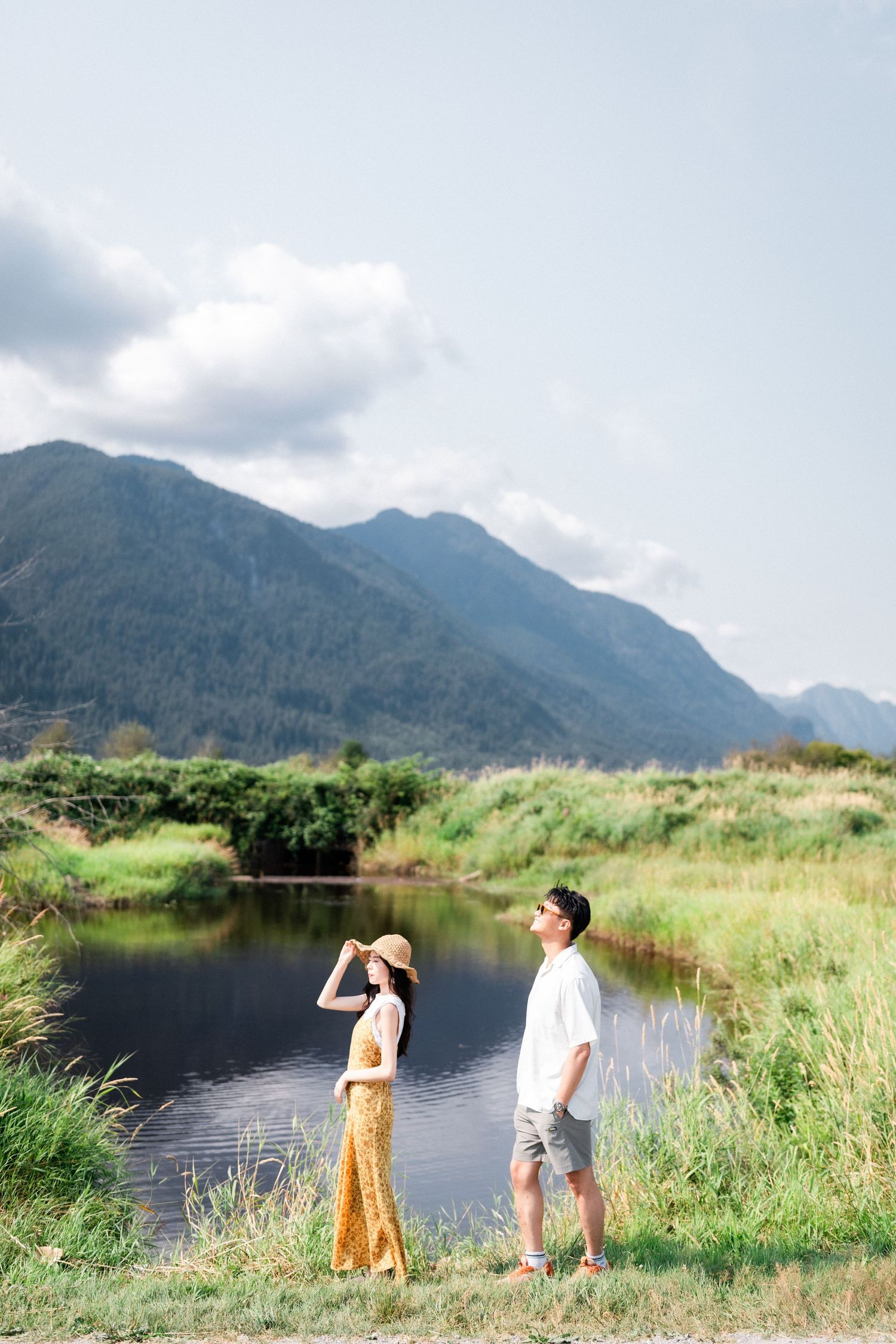 A couple stands by a peaceful pond gazing at dramatic mountain scenery under a partly cloudy sky.