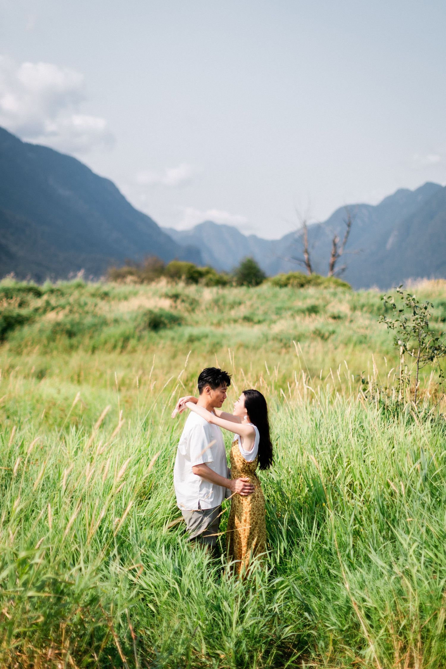 A couple embraces romantically while standing in tall green grass with mountains in the background.