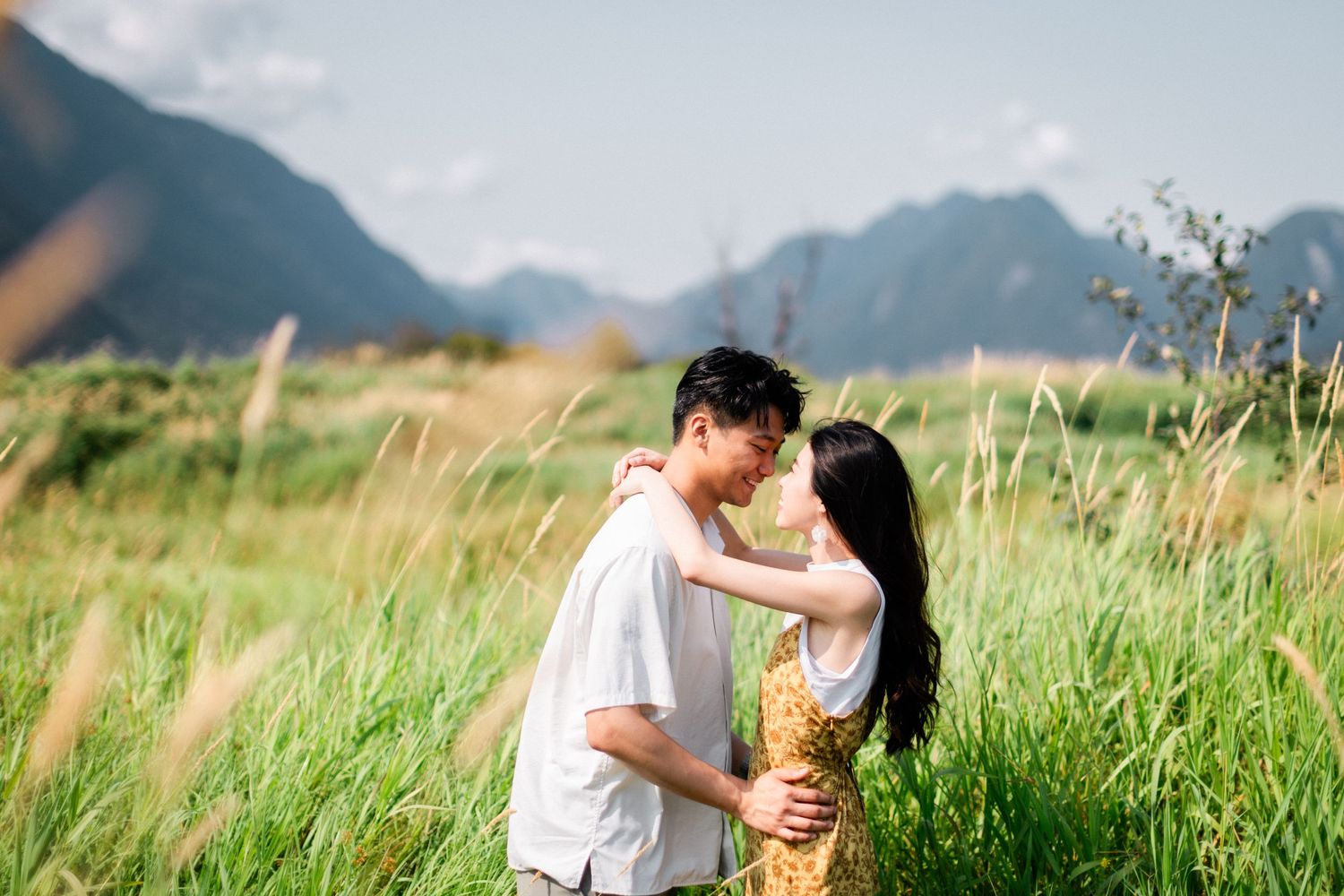 A couple shares a close romantic moment face-to-face in a sunlit green meadow with mountains behind.