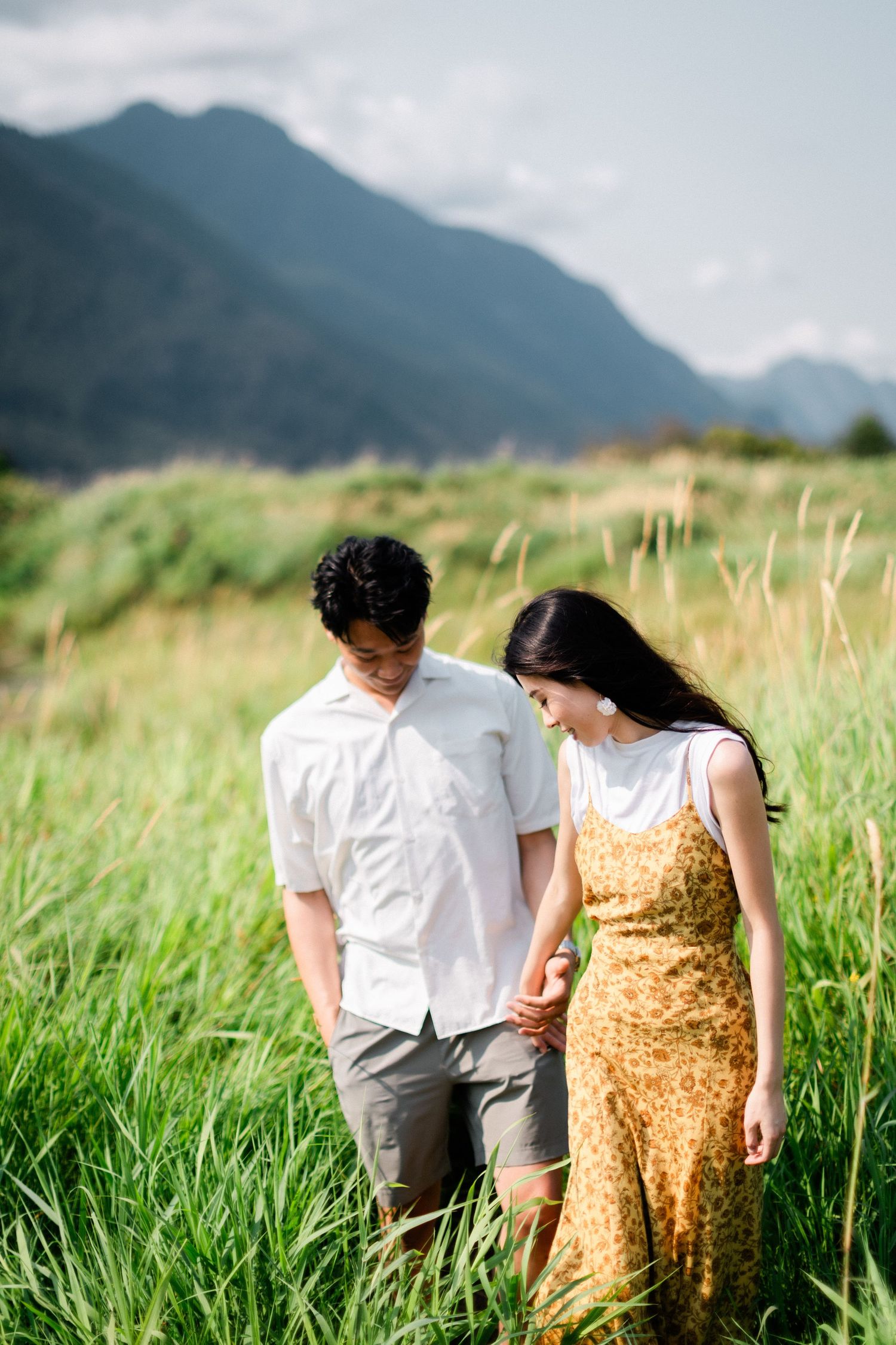 A couple walks hand in hand through tall green grass with scenic mountains visible in the background.
