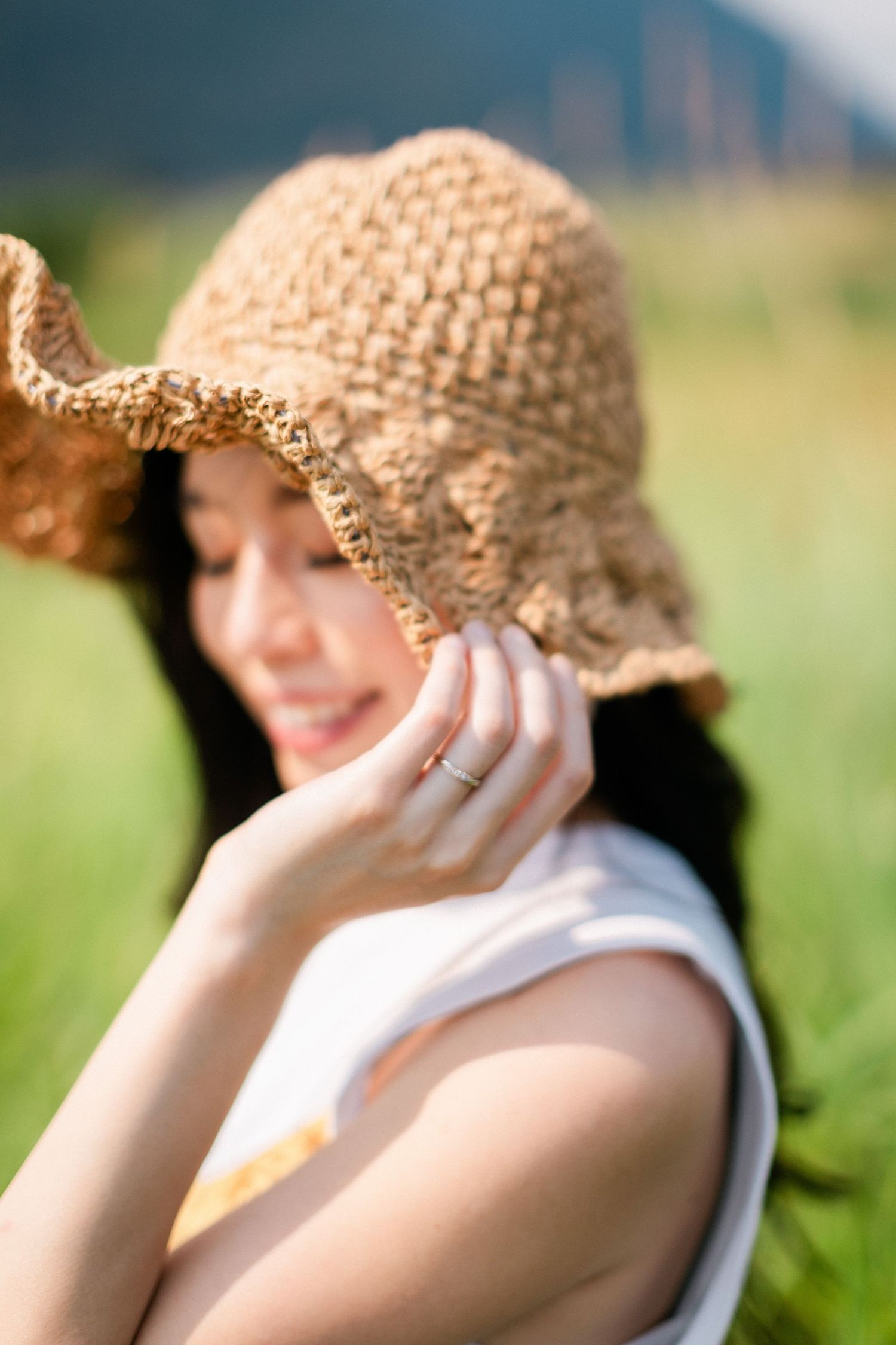 A young woman smiles softly while adjusting her woven straw hat in a sunny outdoor setting.