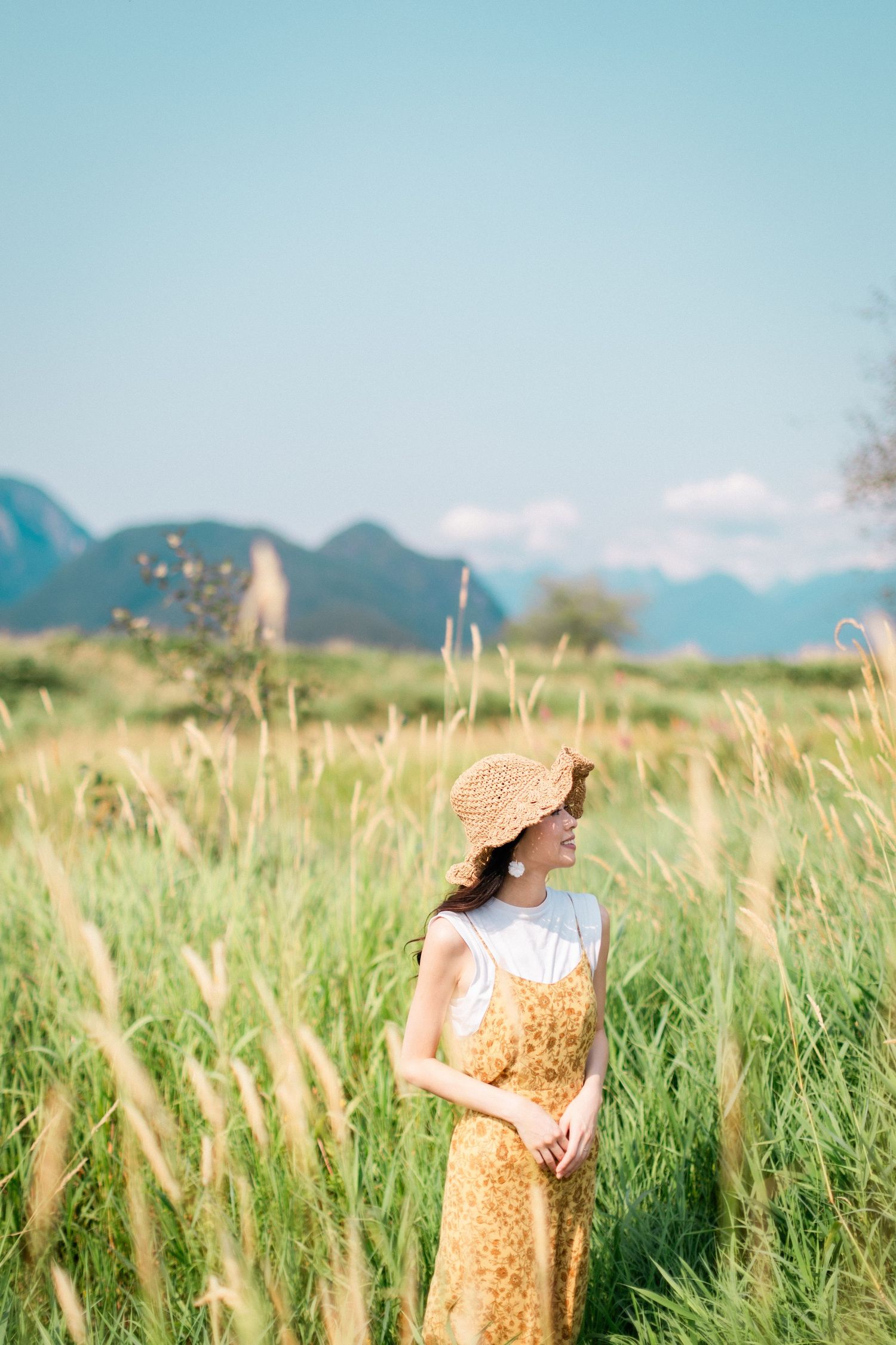 Young woman in yellow floral dress and straw hat standing in tall grass field with mountains in background.