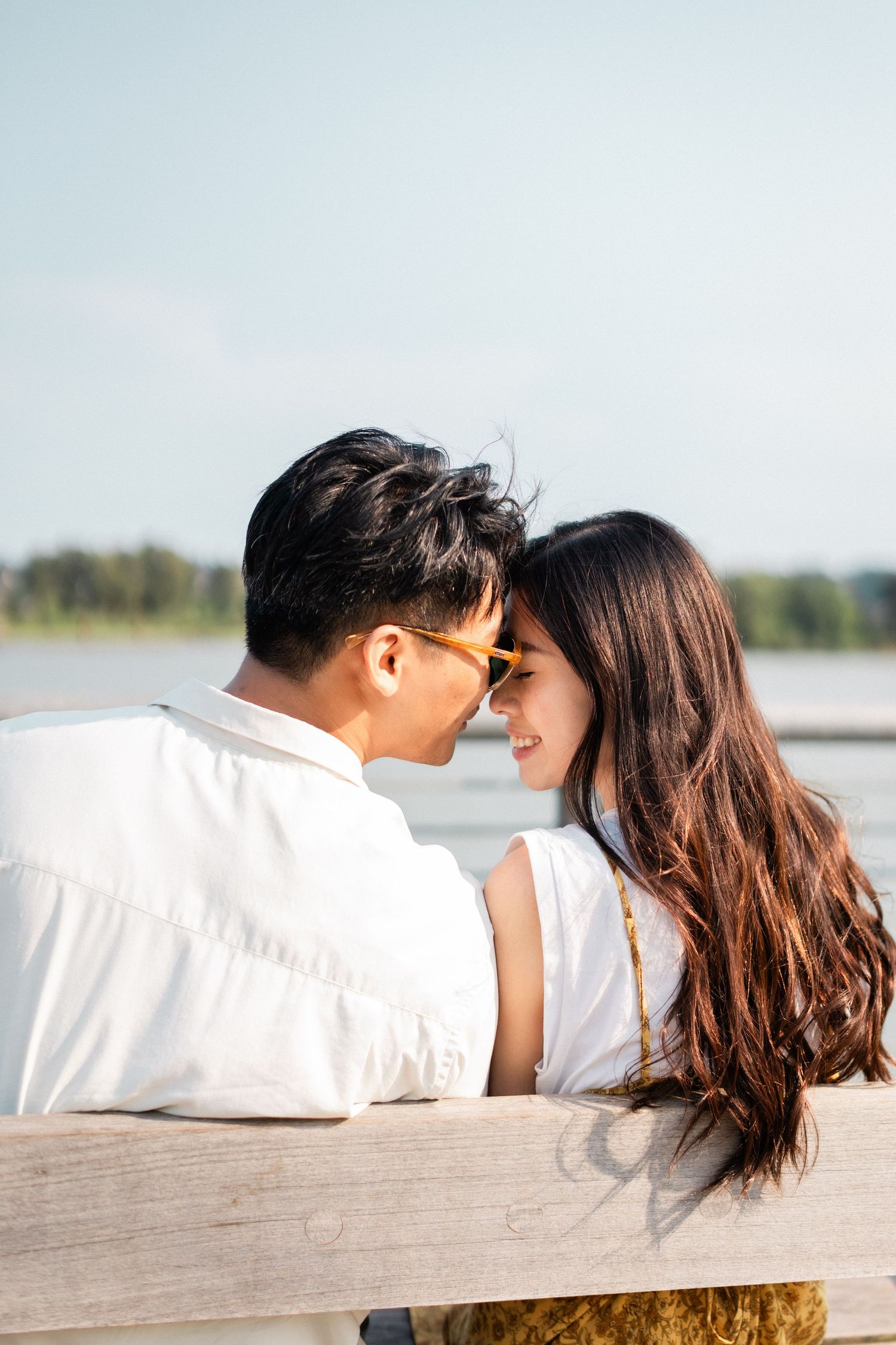 Romantic Asian couple leaning close together on wooden railing by waterfront on sunny day.