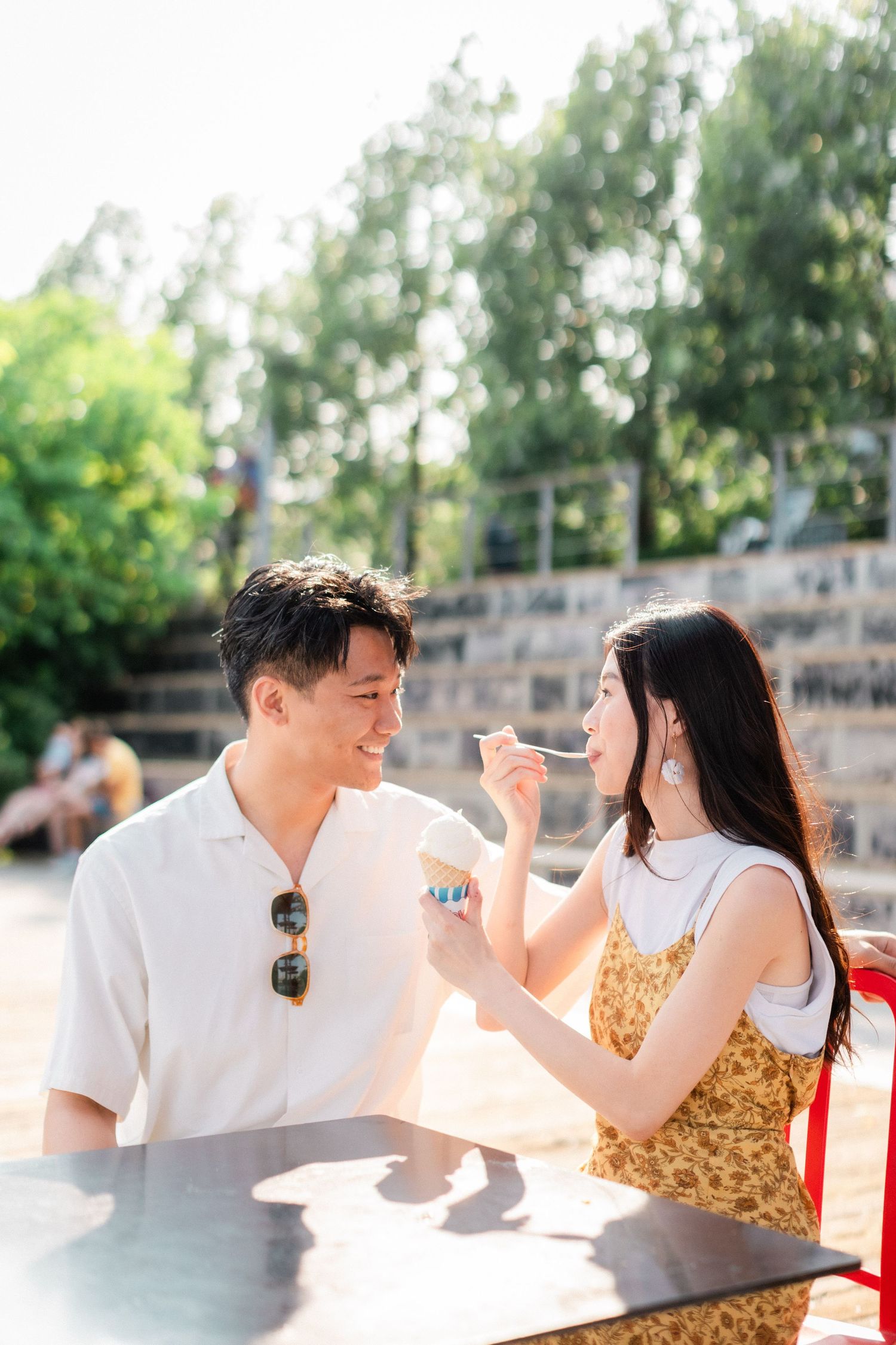 Happy Asian couple sharing ice cream at outdoor cafe table surrounded by green trees.
