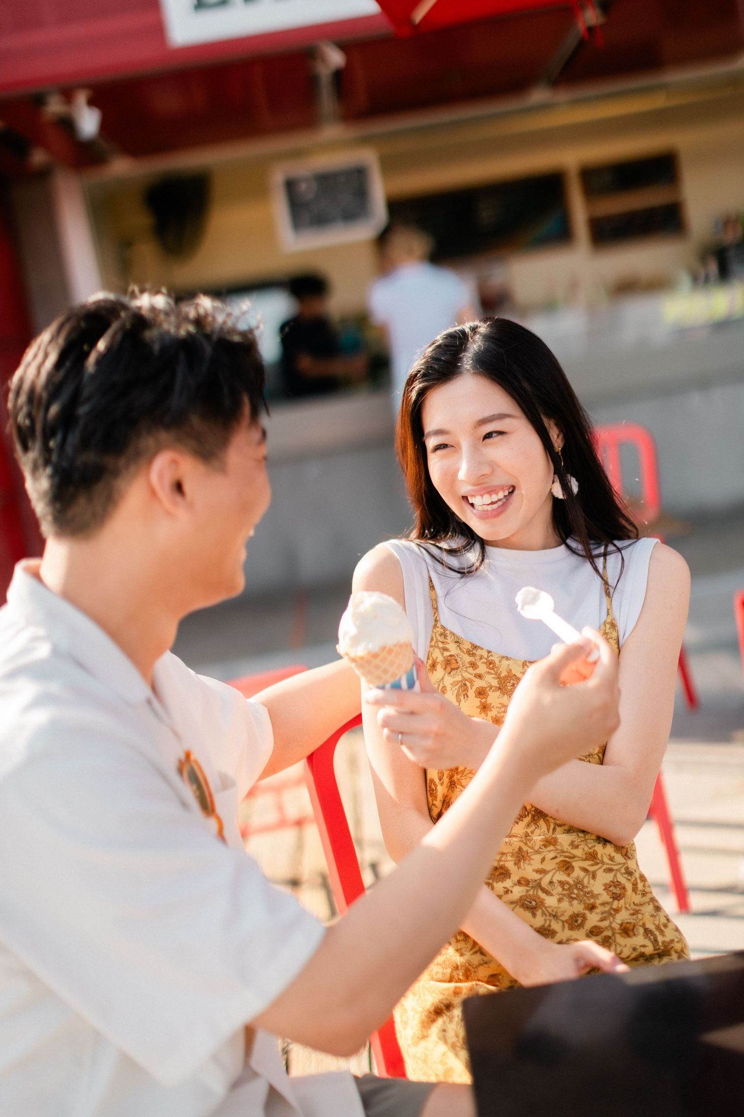 Smiling Asian woman in yellow dress receiving ice cream cone from man at outdoor cafe.