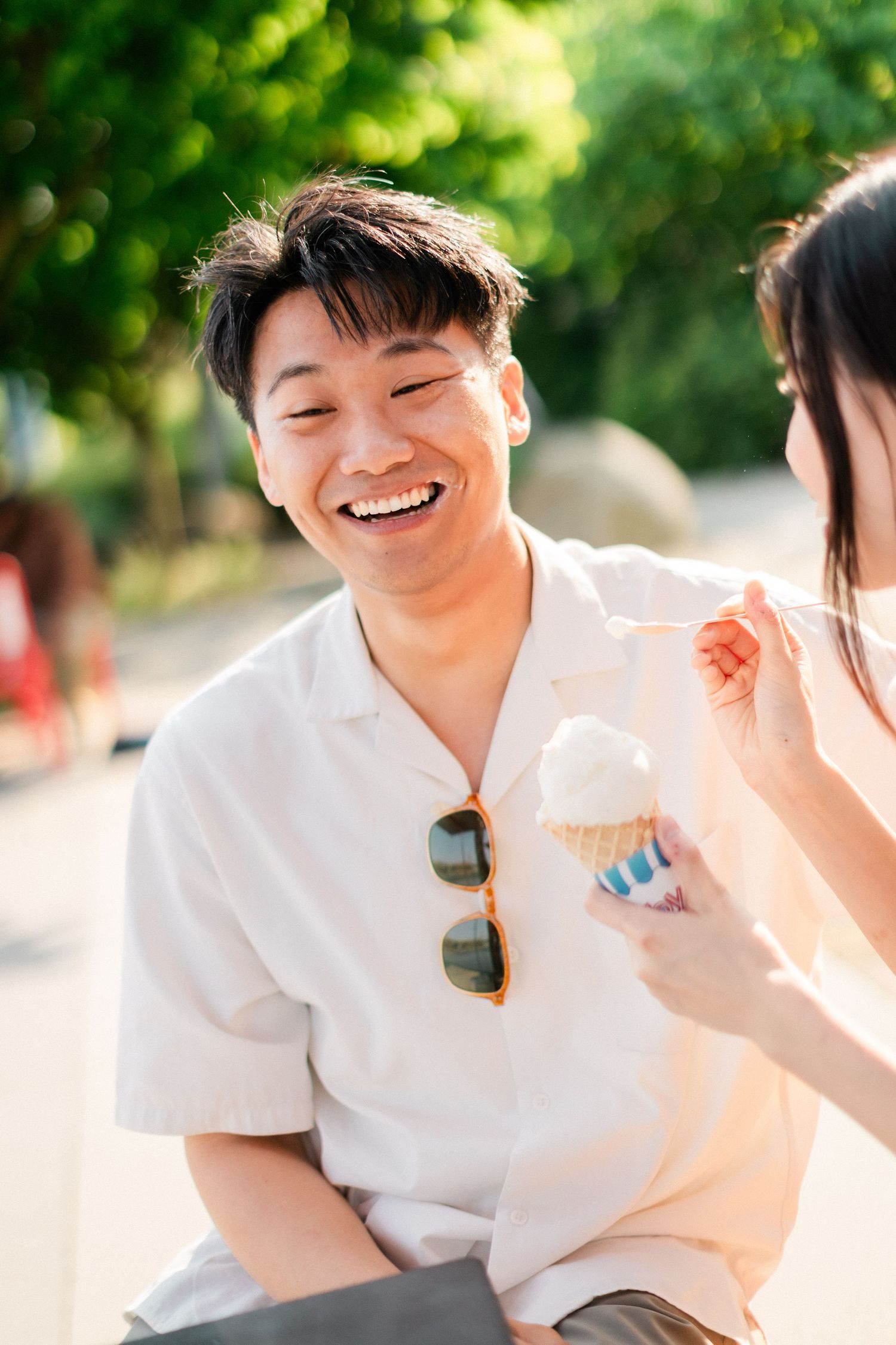 Joyful Asian man laughing while woman feeds him ice cream cone outdoors in sunlight.