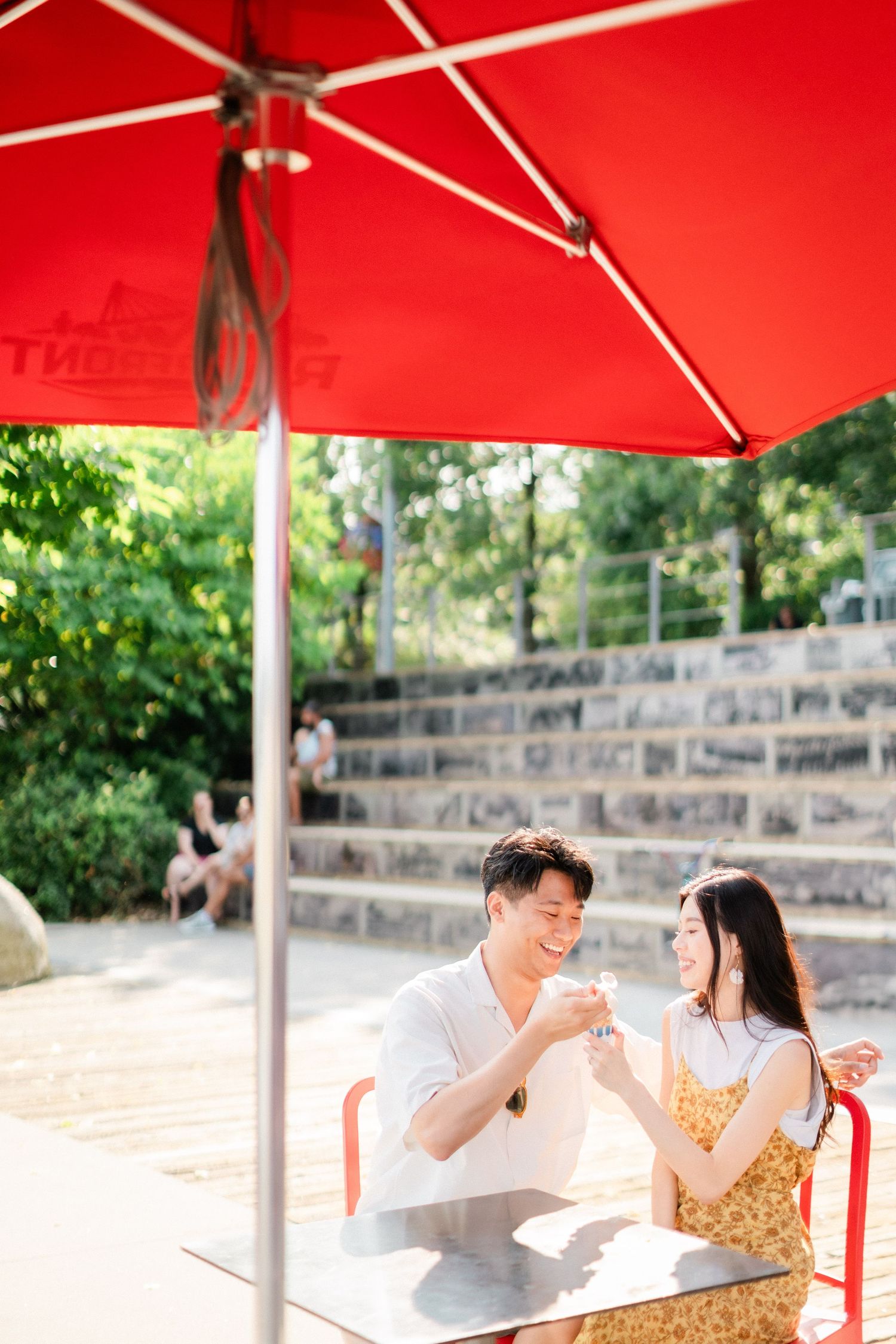 Asian couple enjoying ice cream drinks together under red umbrella at outdoor cafe table.