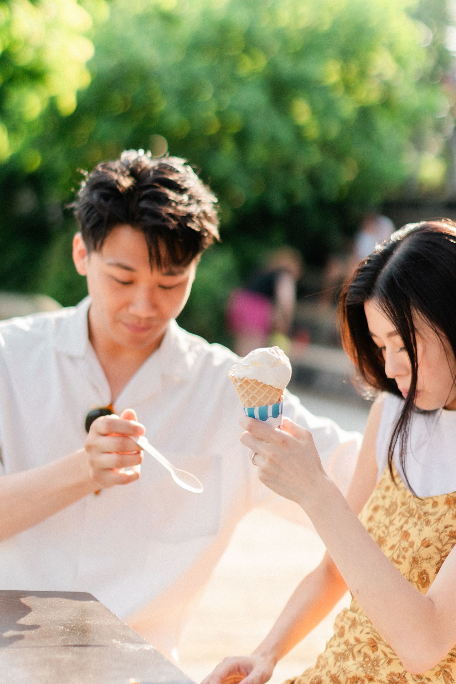Asian couple sharing an ice cream cone together at outdoor table in sunny park setting.